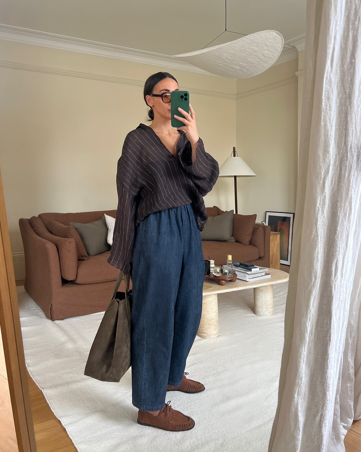 Modelo posa em look estiloso e fácil com camisa oversized listrada, calça azul solta de tecido leve, bolsa marrom e mocassim
