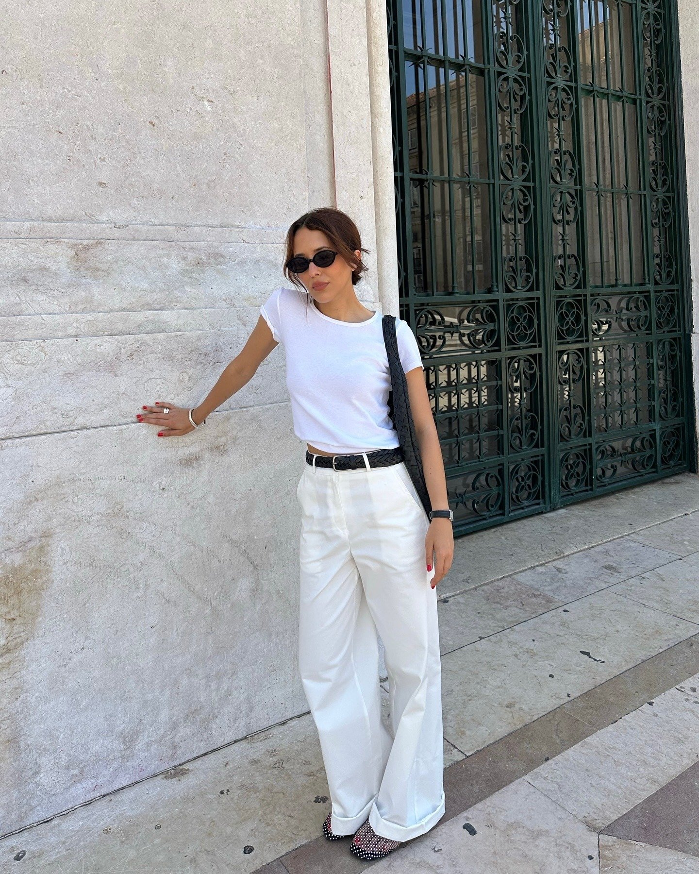 Look estiloso e fácil com camiseta branca justa, calça pantalona off-white, acessórios pretos e postura confiante encostada