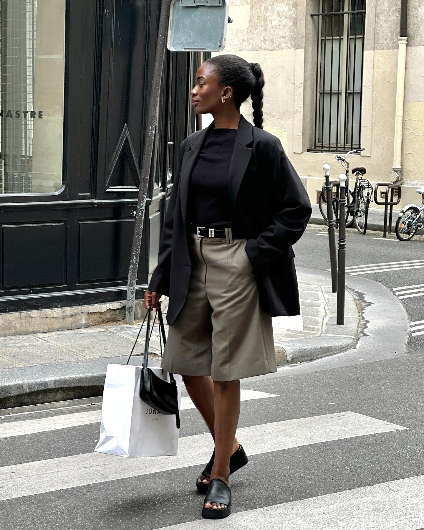 Mulher posa na rua com blazer preto oversized, camiseta preta justa, bermuda bege de alfaiataria e sandália plataforma, look