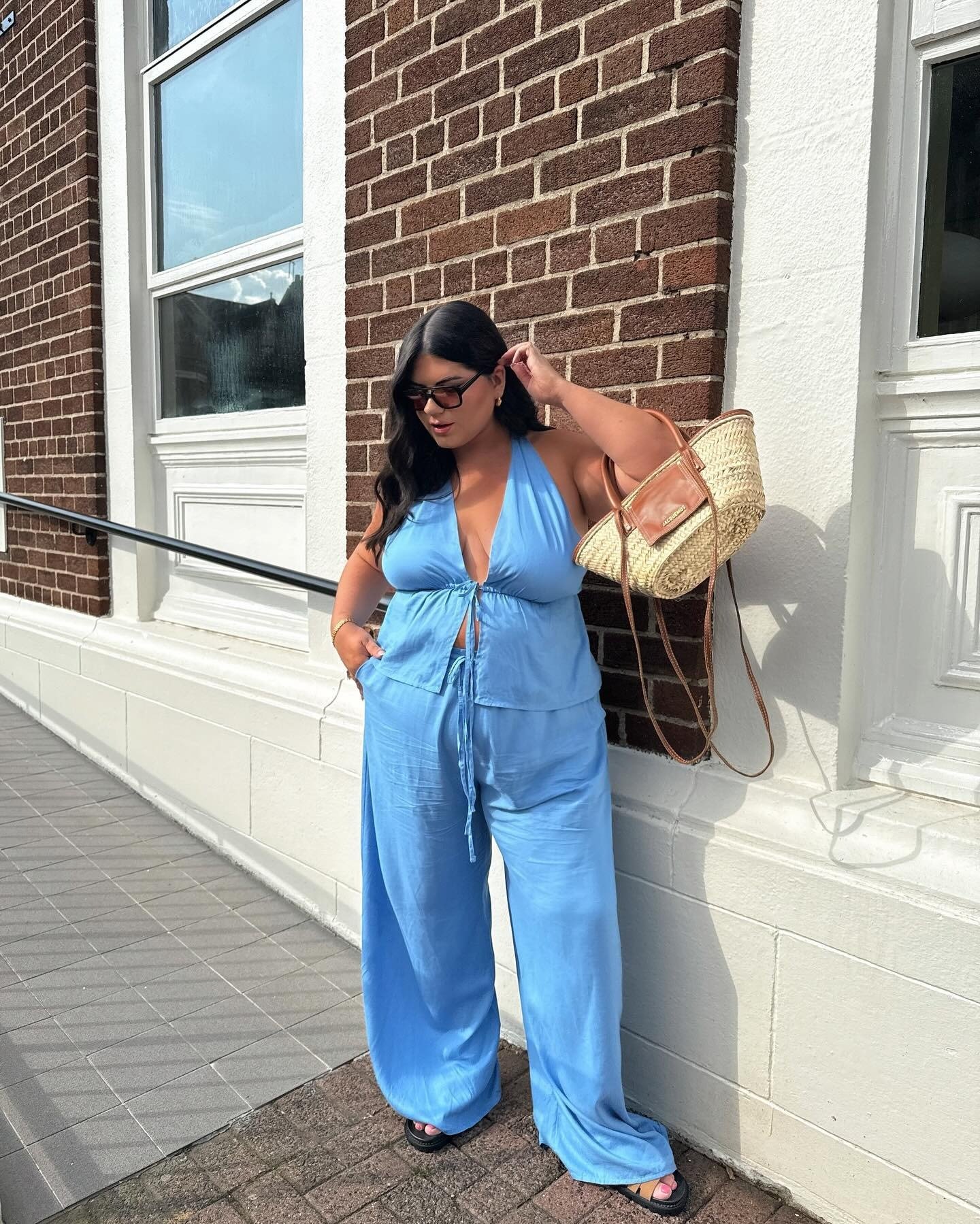 Modelo posa com look estiloso e fácil em conjunto azul de tecido leve, calça ampla e blusa halter, combinando bolsa de palha