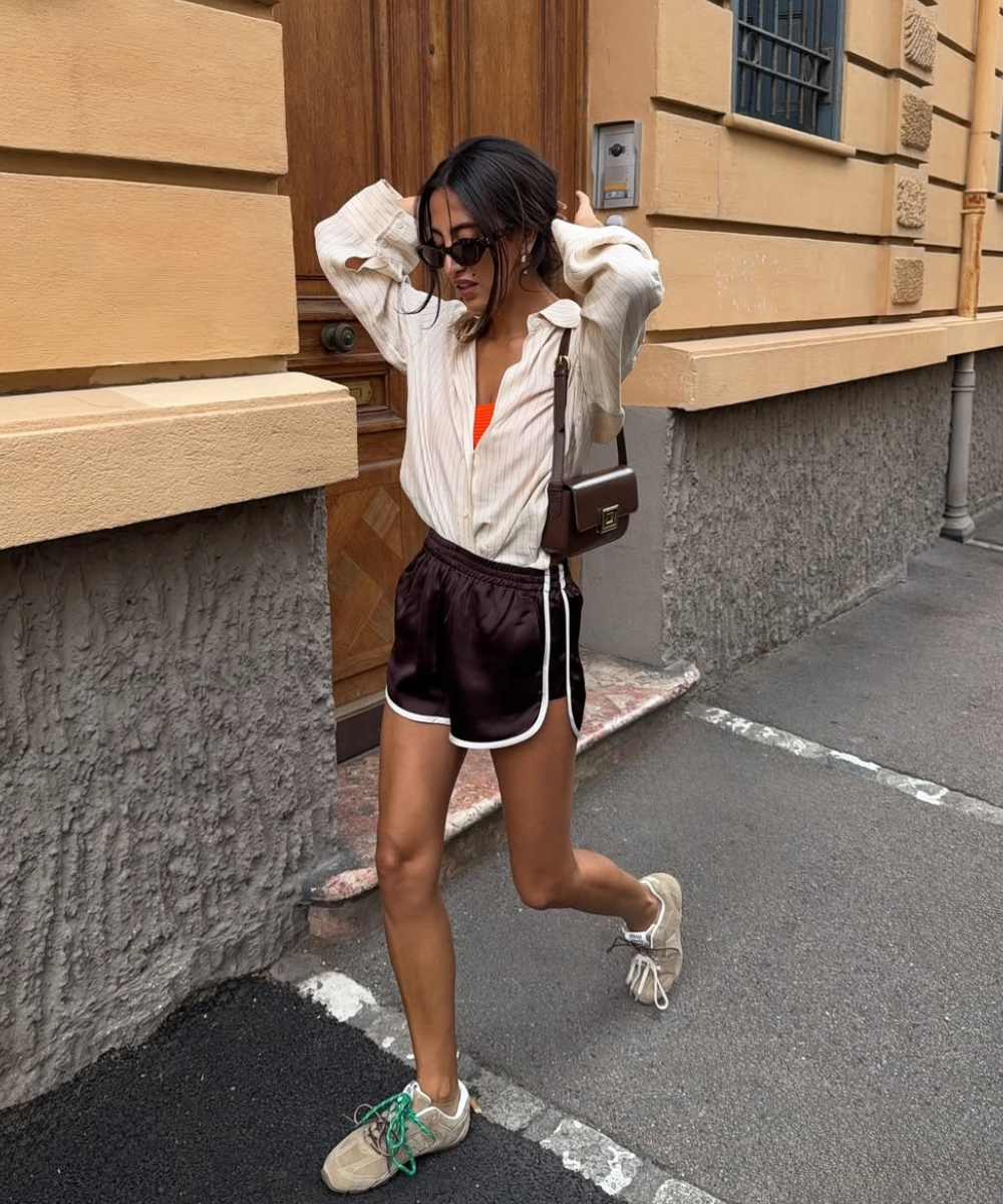 O look apresenta uma combinação estilosa de conforto e modernidade. A pessoa usa uma camisa branca de listras, por dentro de um short esportivo marrom, destacando um top laranja. Os tênis, com detalhes verdes, complementam o visual despojado.