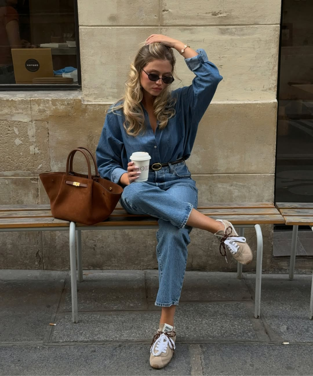 A look fácil com tênis é exemplificado por uma jovem sentada em um banco, usando um conjunto jeans de camisa e calça. Ela segura um copo de café e exibe tênis claros com detalhes em marrom.