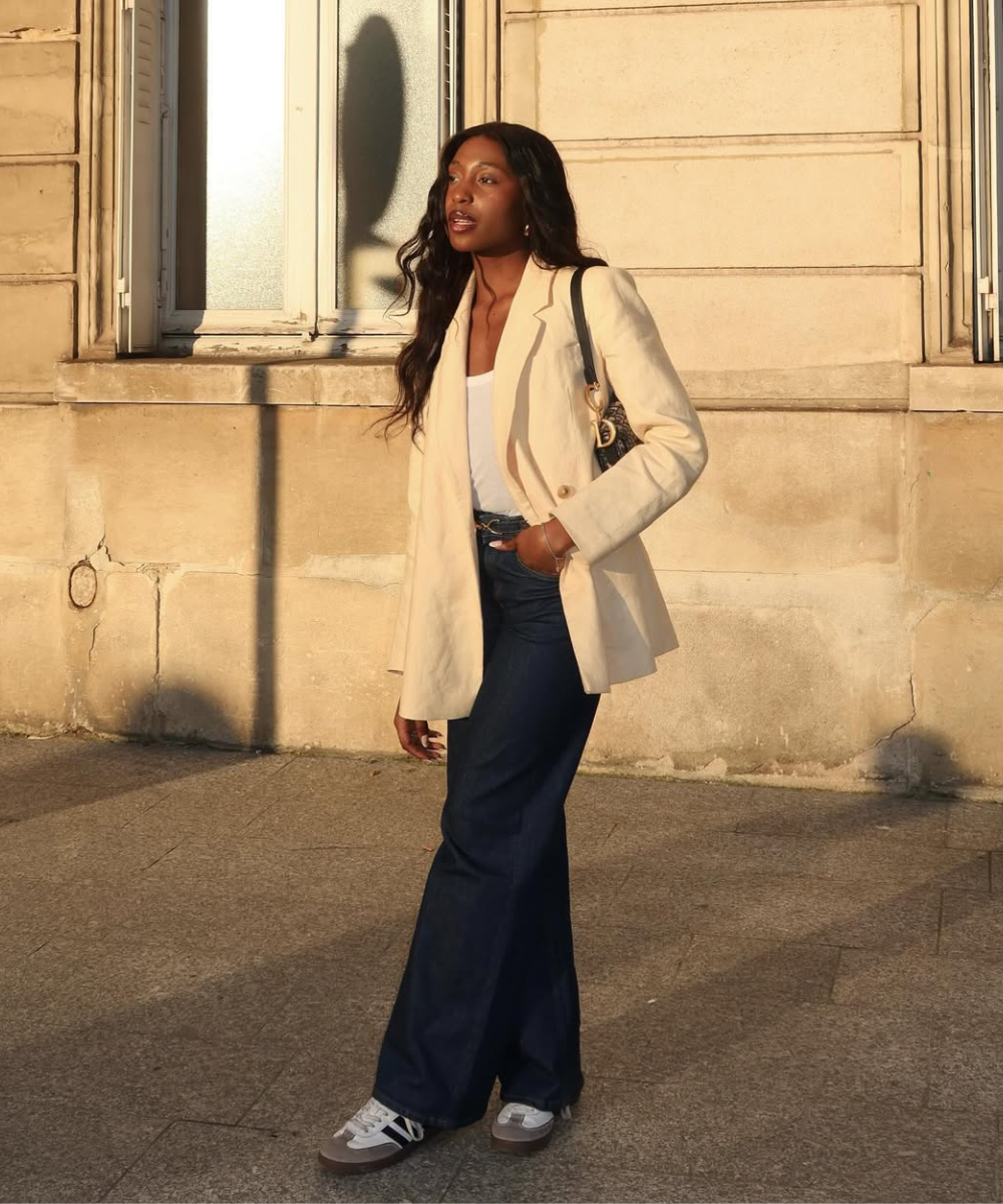 O look apresenta uma combinação estilosa de um blazer claro sobre uma blusa branca, complementado por jeans escuros de corte reto. Os tênis cinzas adicionam um toque casual e confortável, ideal para um visual moderno e prático.