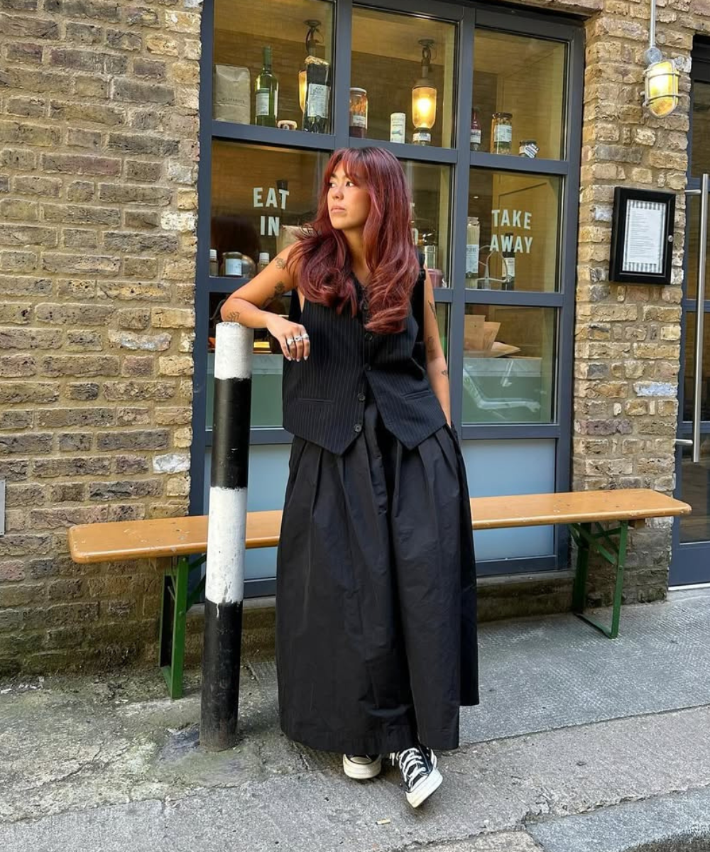 Looks fáceis com tênis: apresenta uma combinação estilosa e descontraída. A modelo usa um colete preto listrado sobre uma saia longa, também preta, criando um visual elegante e moderno. Os tênis brancos adicionam um toque casual, equilibrando o look.