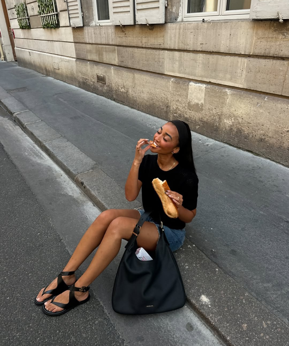 Modelo senta na calçada com look fácil de calor em camiseta preta de malha leve, shorts jeans e sandálias rasteiras pretas,