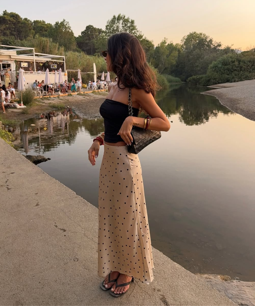 O look apresenta uma blusa preta justa, combinada com uma saia longa e leve em tom bege com poás pretos, ideal para dias quentes. A modelo usa sandálias confortáveis e carrega uma bolsa de corrente.