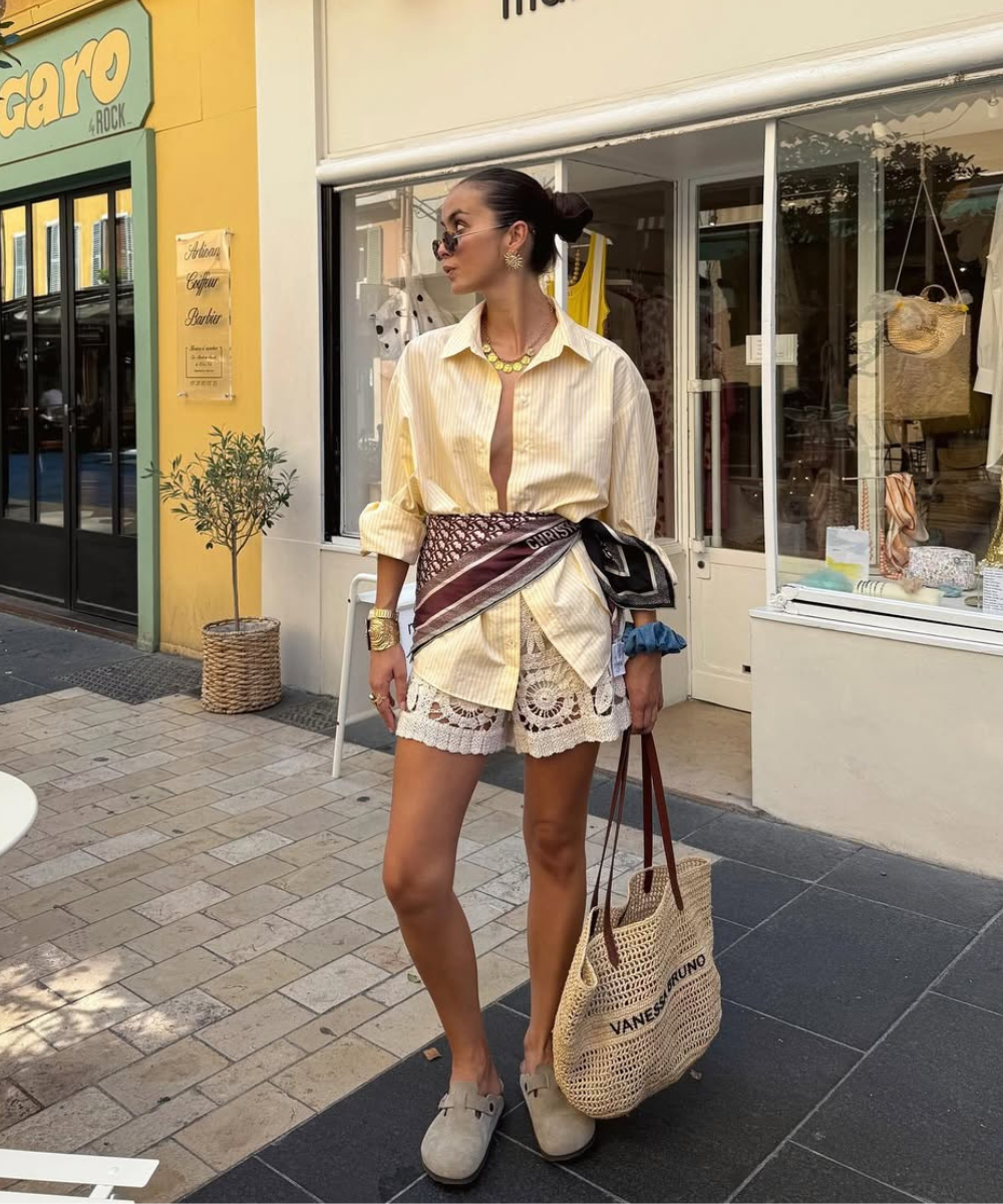 O look é composto por uma camisa de listras em tons claros, com botões abertos, que confere um ar descontraído. A cintura é marcada por um cinto estiloso e um lenço, que traz um toque de sofisticação.