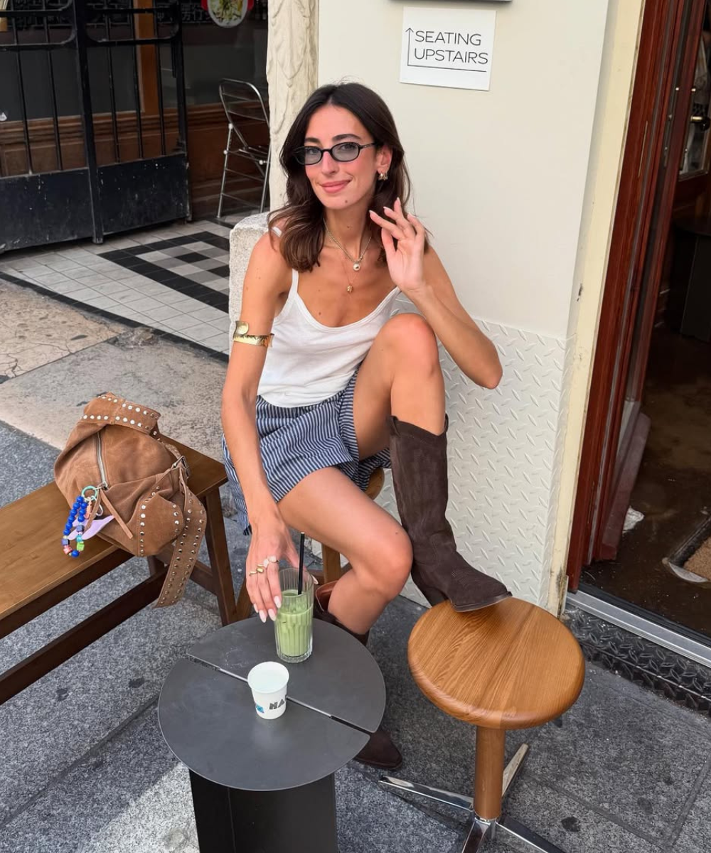 Look fácil e fresquinho, perfeito para dias quentes. A modelo veste uma regata branca e uma saia listrada, combinando conforto e estilo. As botas de camurça e os acessórios discretos, como anéis e pulseira, complementam o visual.