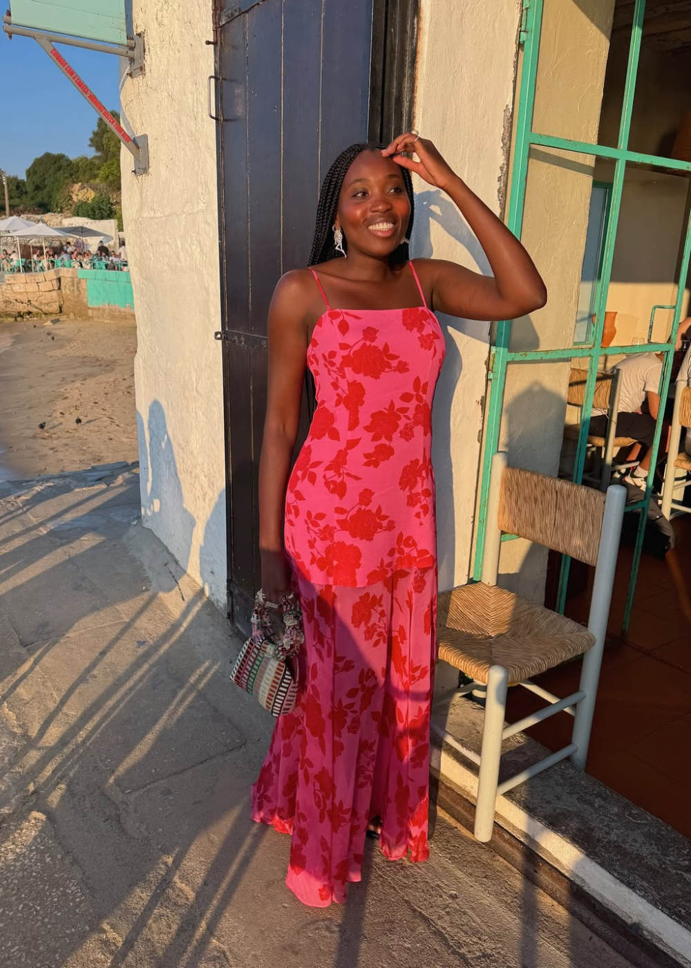 Modelo sorri e posa com vestido longo rosa de alças finas e estampa floral vermelha, em clima leve e ensolarado, ideal para