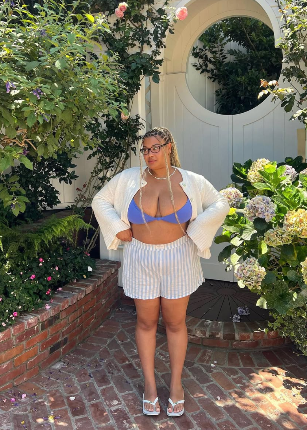 Modelo posa confiante com look para o verão composto por top azul, shorts listrados de algodão e cardigan branco leve.