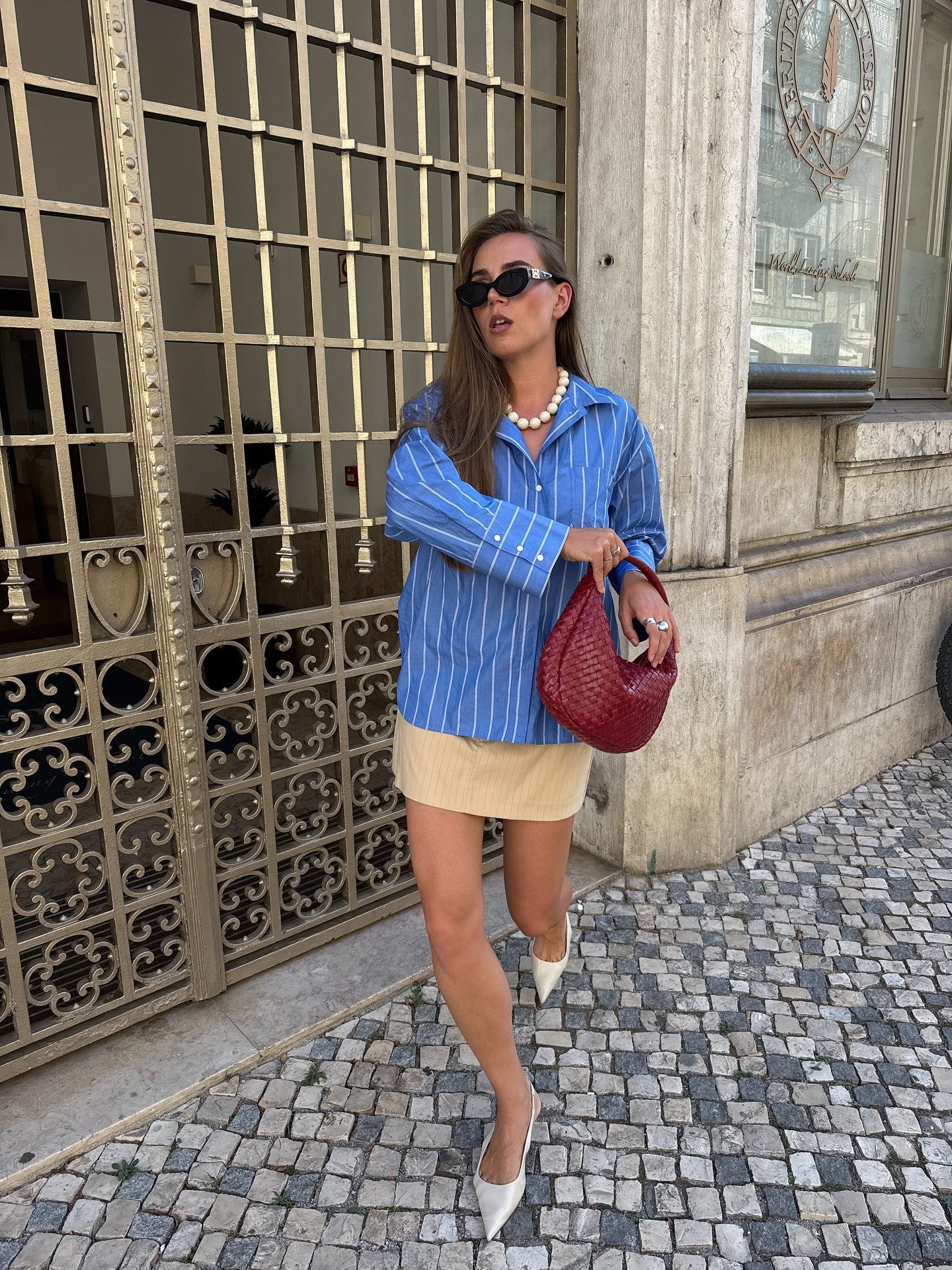 Modelo posa com camisa azul listrada oversized, saia bege curta, scarpin branco. Ideias de looks femininos.