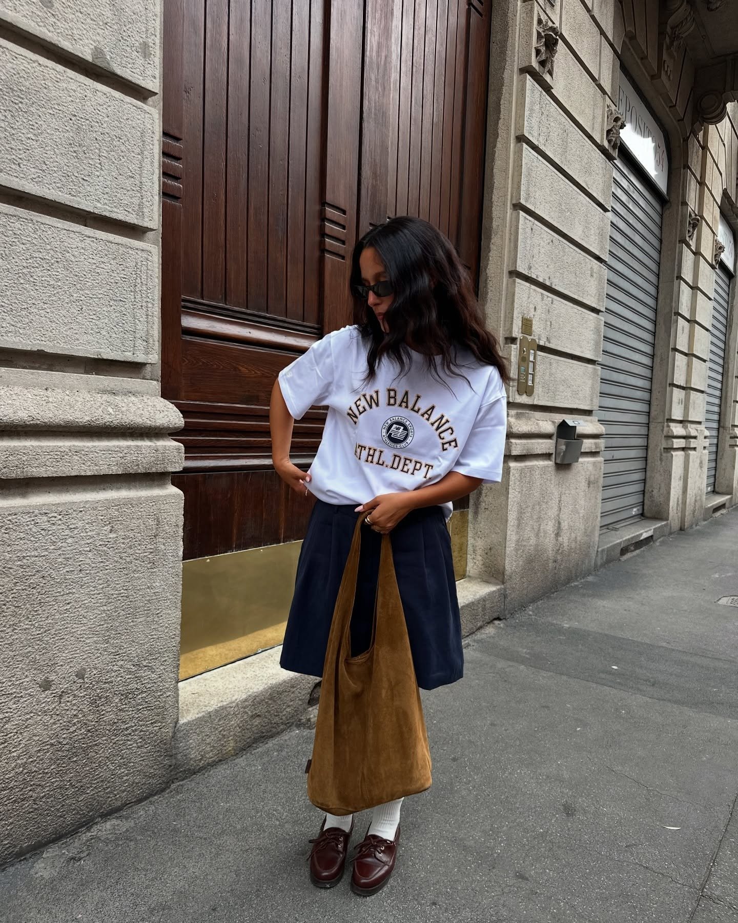 Ideias de looks femininos com camiseta branca oversized, saia azul escura plissada, bolsa de camurça marrom e mocassim vinho.