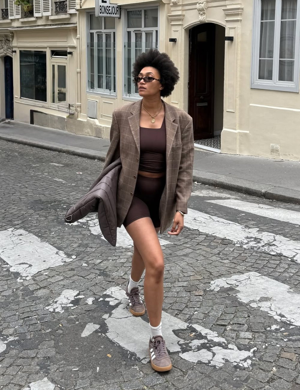 A imagem apresenta uma mulher caminhando com estilo urbano, vestindo um short marrom e tênis em um tom neutro. Ela combina o look com um blazer xadrez e uma blusa cropped, criando um contraste interessante entre o casual e o sofisticado.