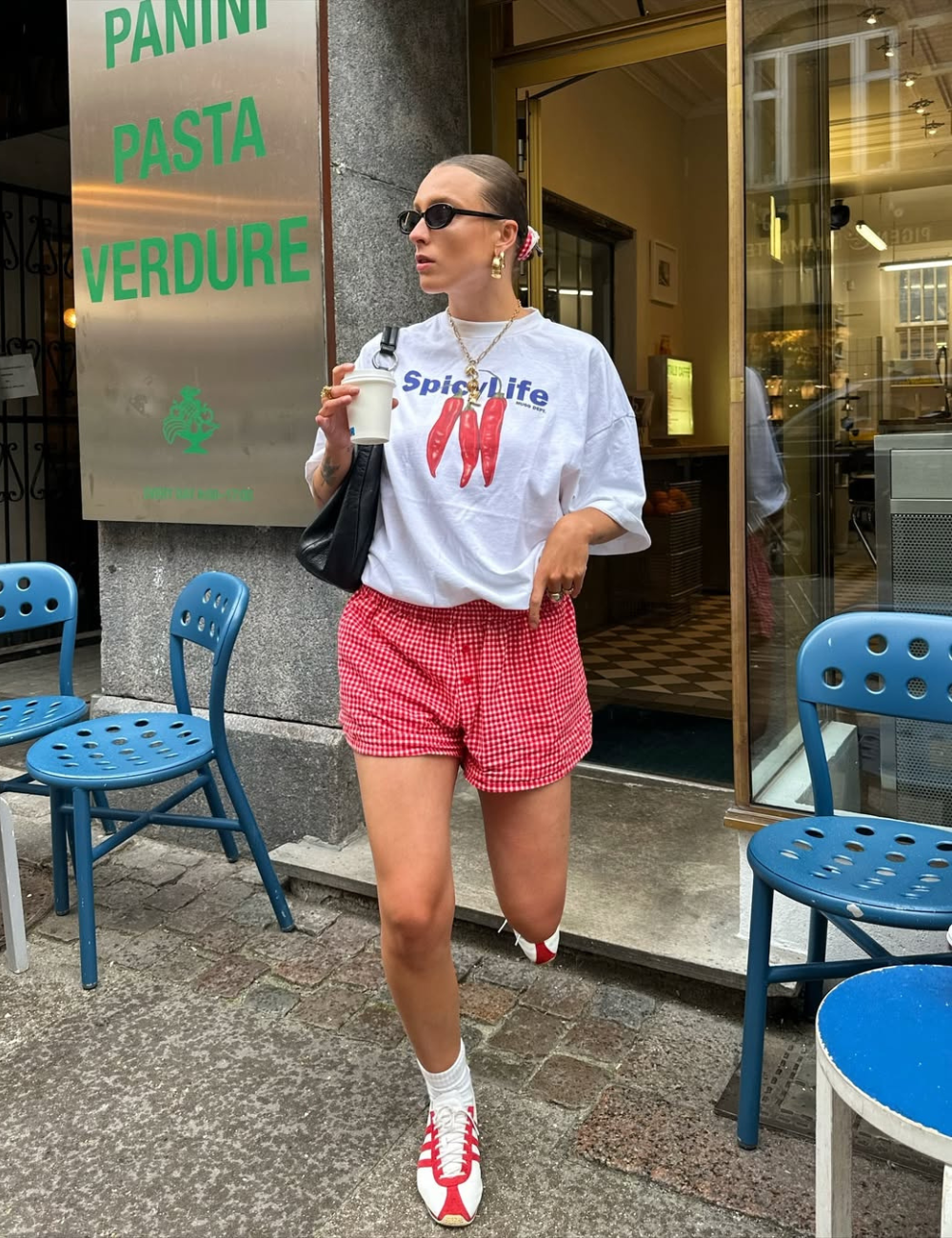 A combinação de short vermelho de estampa xadrez com uma camiseta branca oversized traz um ar descontraído e moderno. O look é complementado por tênis brancos com detalhes vermelhos e meias brancas, criando um contraste estiloso.