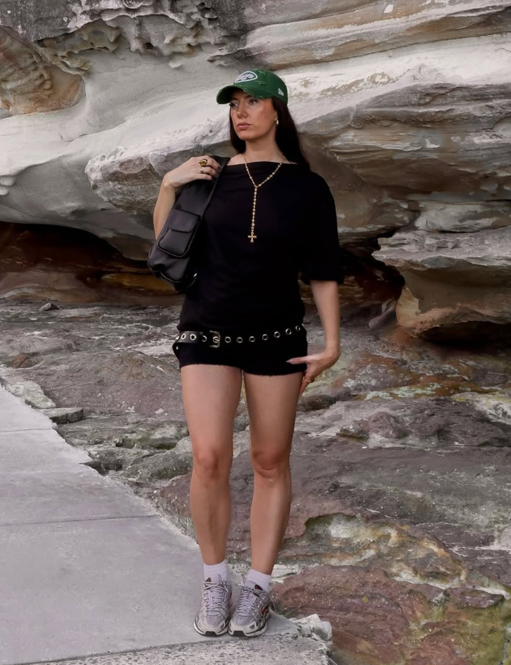 A imagem apresenta uma mulher estilosa vestindo um short preto com detalhes em fivela, combinado com uma camiseta preta e tênis confortáveis. Ela usa um boné verde, que complementa o look casual e moderno.