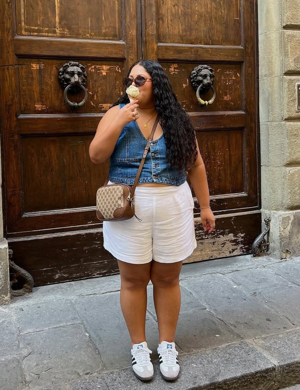A imagem apresenta uma mulher usando um short branco e tênis, combinados com um colete jeans e óculos escuros, criando um look casual e moderno. Ela segura um sorvete, destacando um estilo descontraído, ideal para dias quentes.