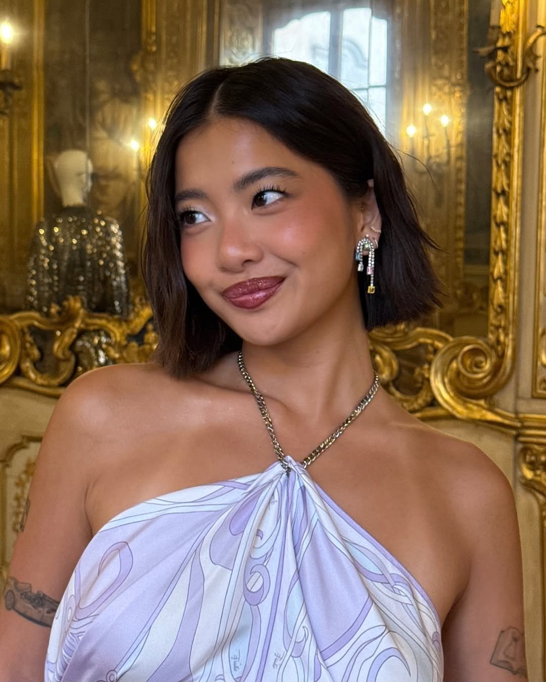 A imagem apresenta uma jovem com pele natural feita com melhores iluminadores para uma maquiagem glow. Ela usa um vestido com estampa suave e um colar delicado. O ambiente luxuoso, com detalhes em dourado e luz suave, complementa o look.