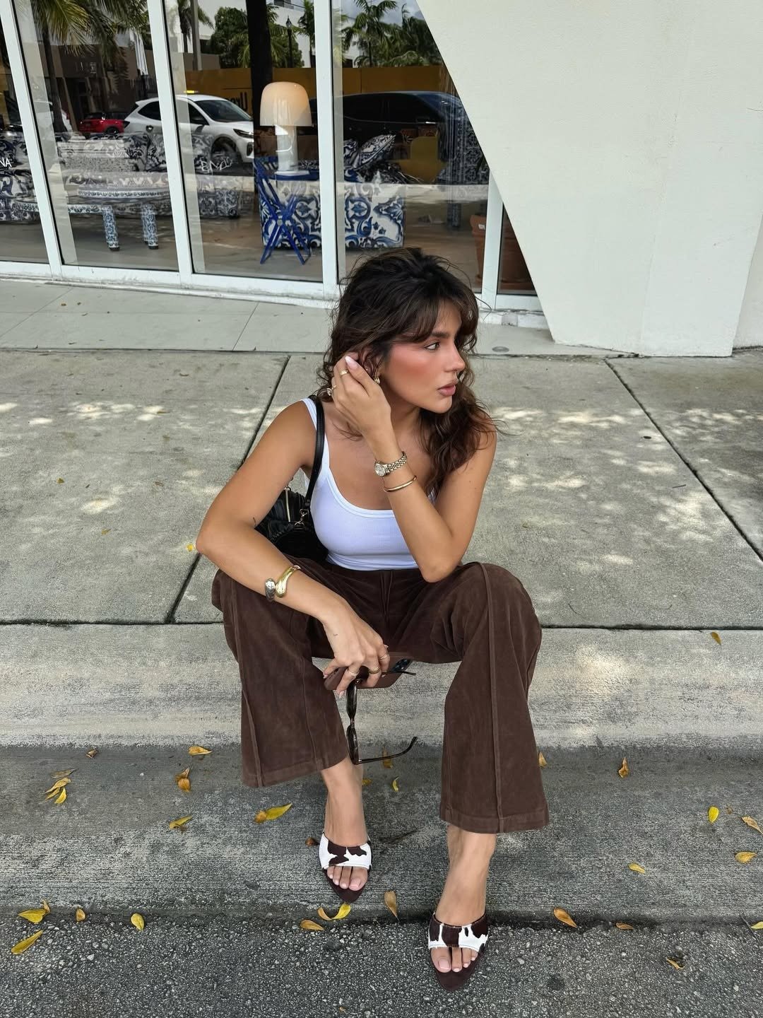 Uma jovem senta-se em uma calçada, usando uma blusa branca e calças marrons de tecido leve. Seu cabelo solto e levemente ondulado emoldura o rosto, que exibe um iluminador sutil, feito com os melhores iluminadores para uma maquiagem glow