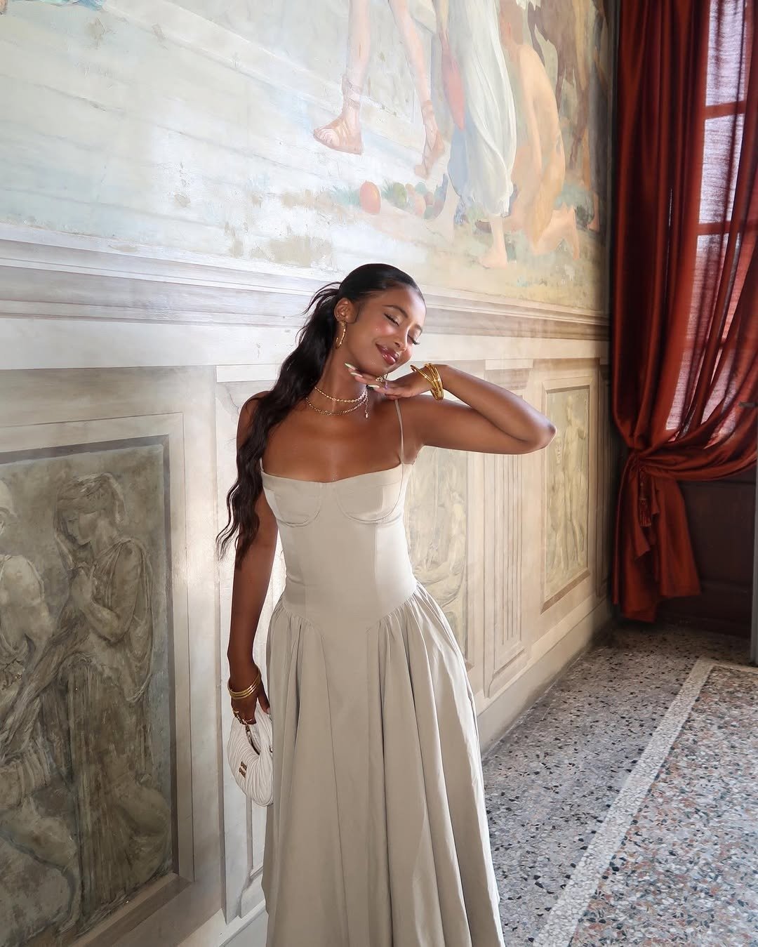 Uma mulher com vestido longo e fluido na cor bege, cabelo está solto e levemente ondulado, complementado por acessórios delicados, como colares e pulseiras douradas e uma pele feita com os melhores iluminadores para uma maquiagem glow