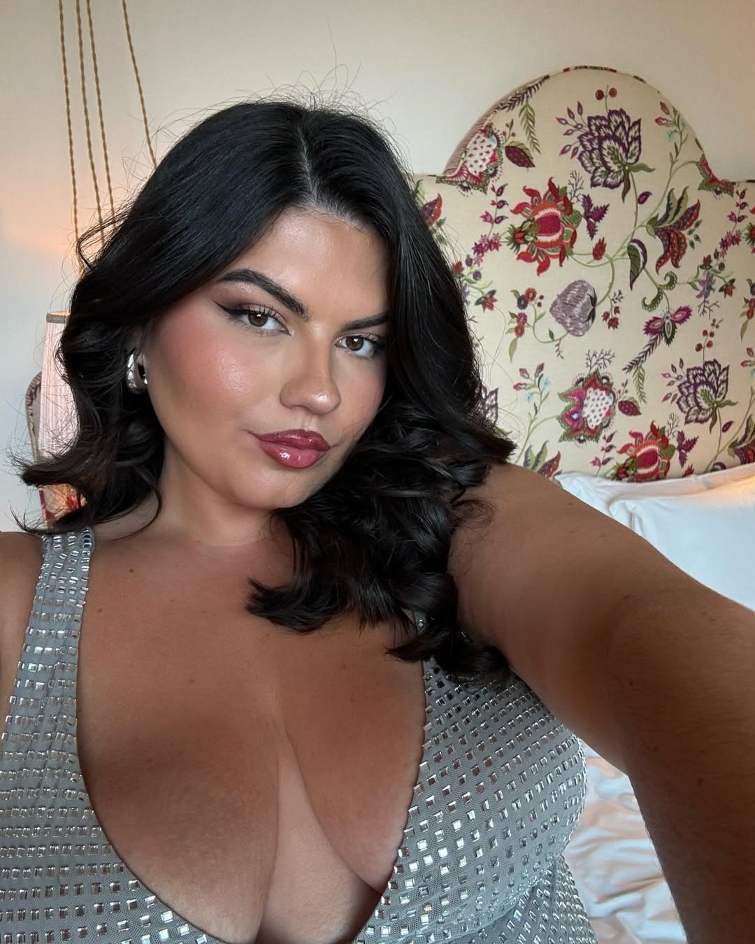 A imagem apresenta uma mulher com cabelo solto e ondas suaves, usando um vestido prateado com detalhes brilhantes. Sua maquiagem destaca um glow natural, com os melhores iluminadores para uma maquiagem glow de verão