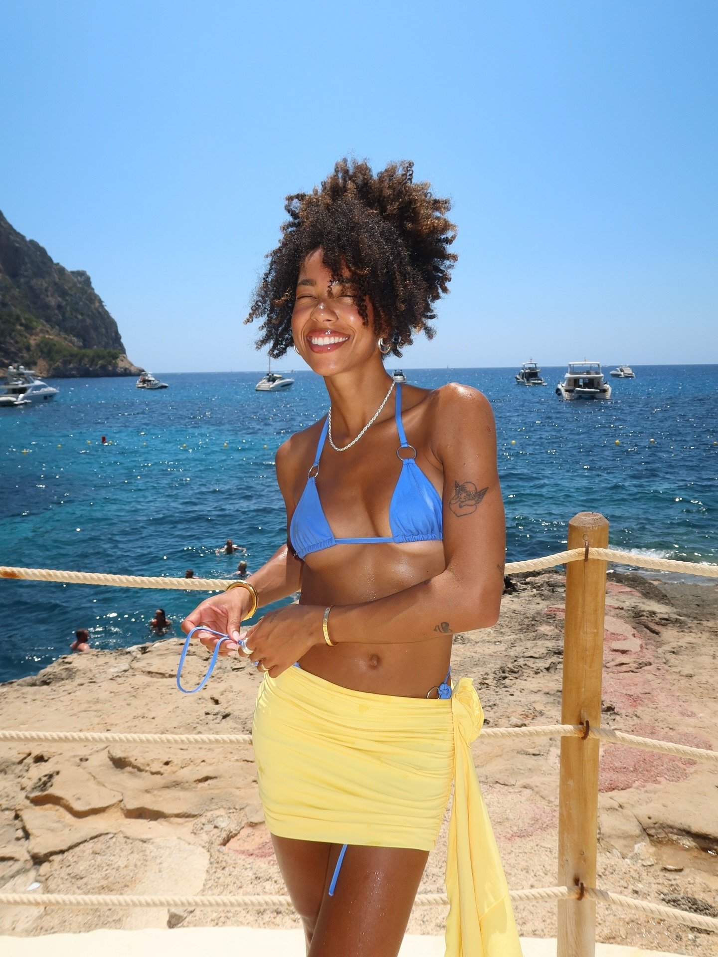 Modelo sorri com maquiagem para praia leve, usando biquíni azul e saia amarela de tecido fluido, em cenário ensolarado à