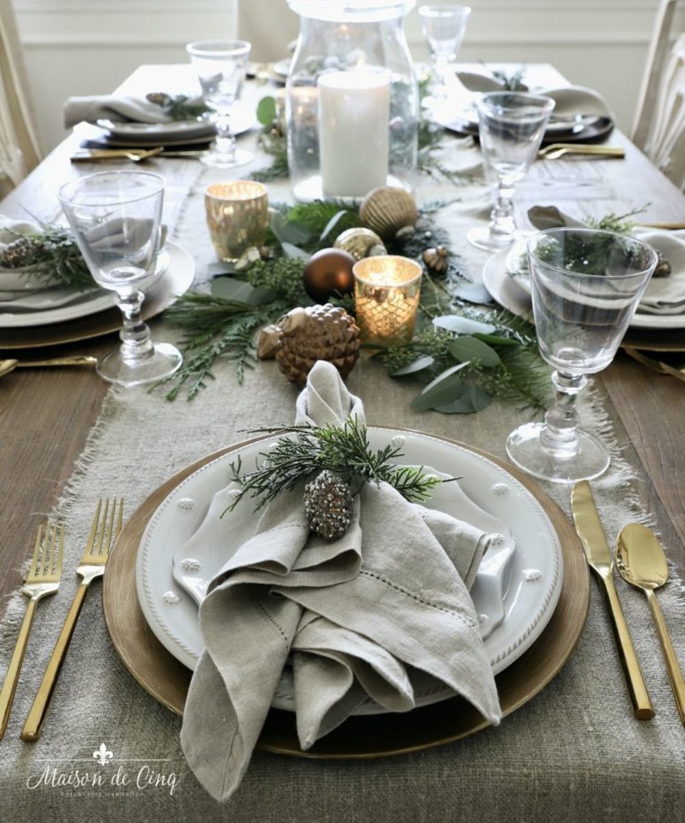 Mesa de natal simples e bonita com louças brancas, talheres dourados, guardanapos de linho, folhagens verdes e velas.