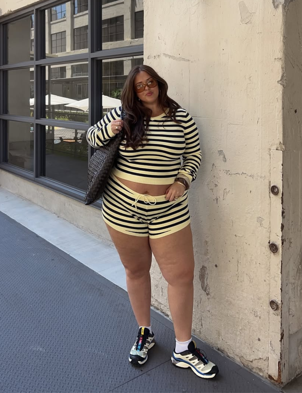 Look com micro shorts em listras amarelas e pretas, combinando com um cropped de manga longa. A modelo usa tênis esportivos estilizados e carrega uma bolsa grande de palha. Óculos de sol completam o visual moderno e casual, ideal para dias quentes.