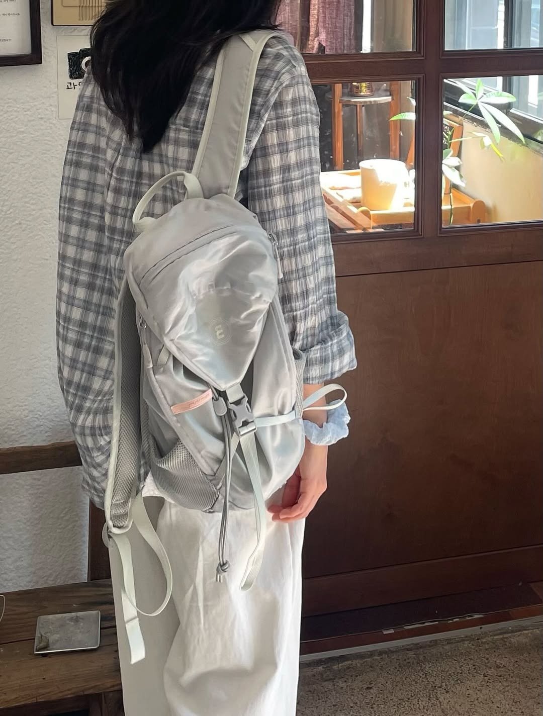 Modelo de costas com postura relaxada usa mochila para viagem cinza clara em tecido leve, combinando com camisa xadrez e