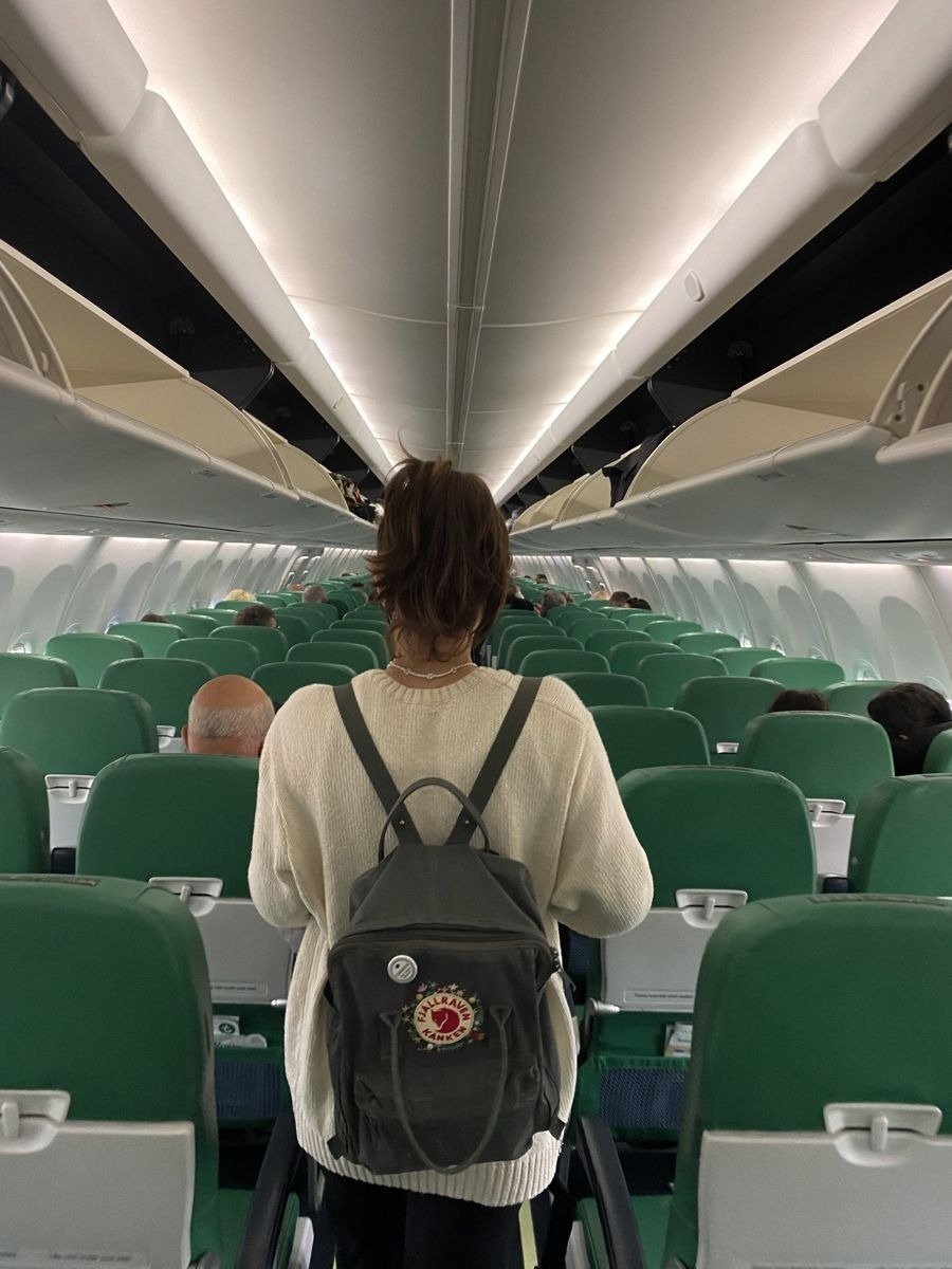 Modelo usa suéter bege e mochila para viagem cinza escuro de tecido resistente com logo vermelho, em cabine de avião com