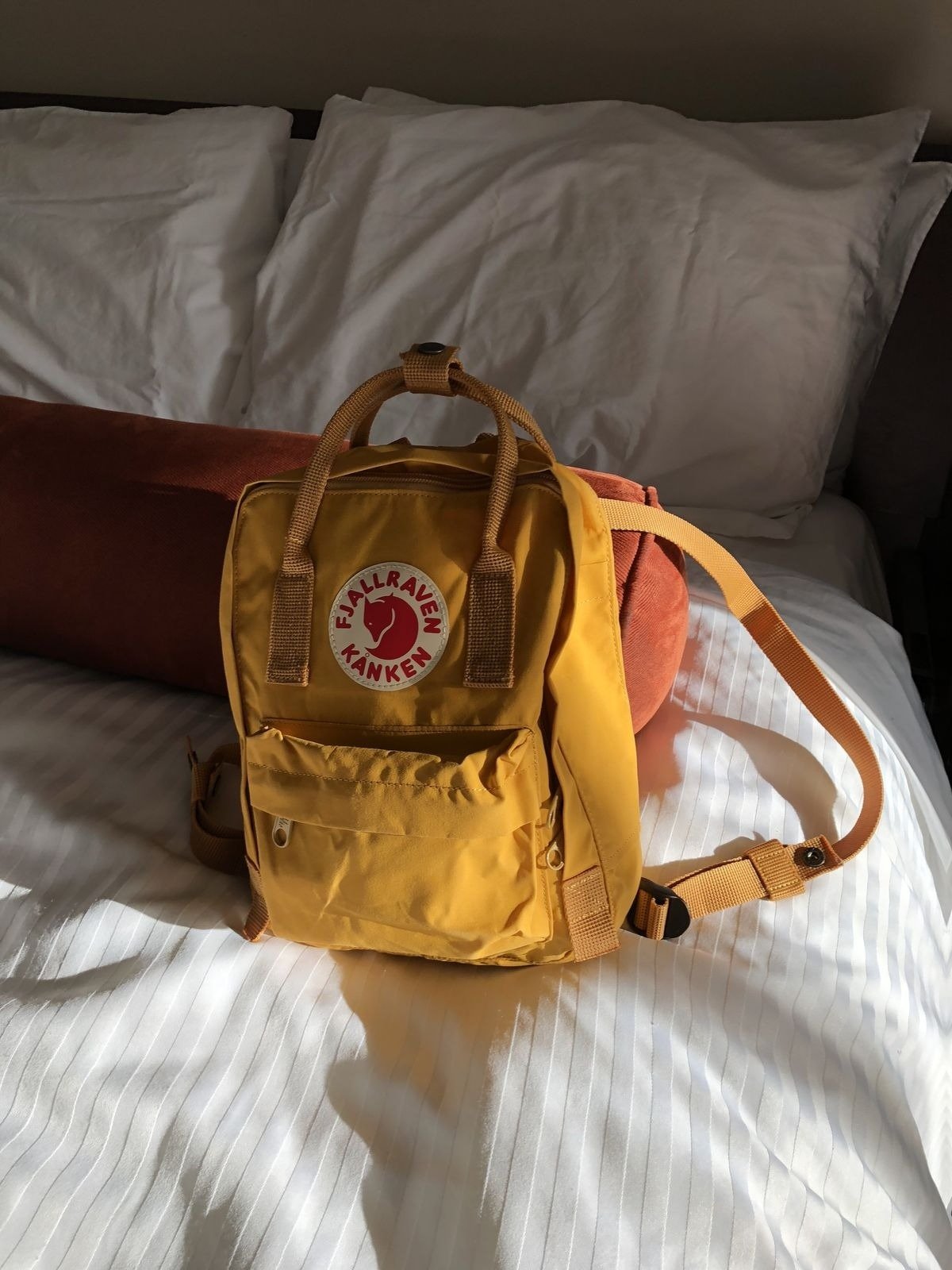 Mochila para viagem amarela em nylon resistente com design compacto e alças ajustáveis, sobre cama branca iluminada por luz