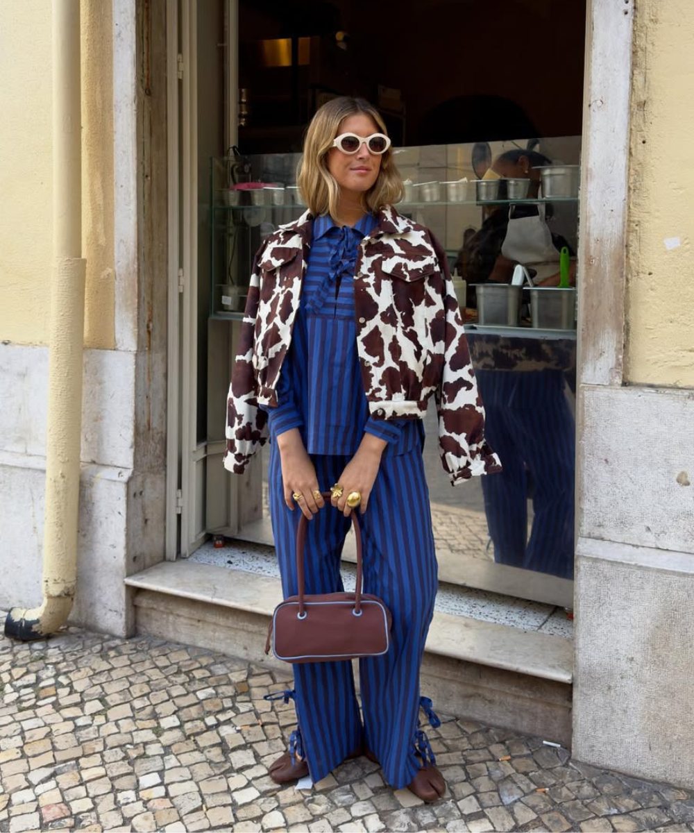 Estilo ousado para outono/inverno 2026: jaqueta de estampa de vaca, blusa listrada azul e calça combinando. Acessórios em ouro e bolsa marrom completam o visual. Óculos brancos destacam o look moderno e arrojado.