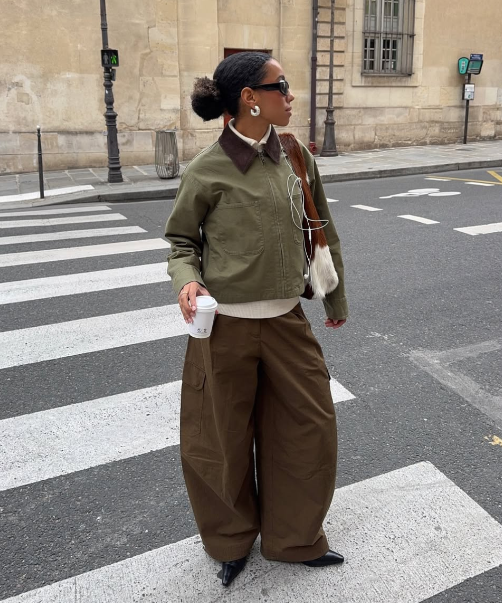 Look moderno e casual para o outono/inverno 2026, com jaqueta verde e calças largas. Acessórios como óculos escuros e brincos grandes complementam o visual. O uso de camadas e texturas é ideal para a estação.