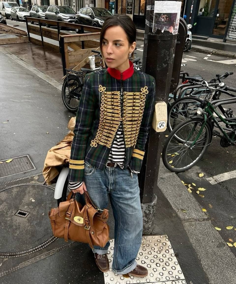 A imagem retrata uma mulher com um estilo ousado para o outono/inverno 2026. Ela usa um blazer xadrez com detalhes dourados e uma gola vermelha, combinado com uma blusa listrada.