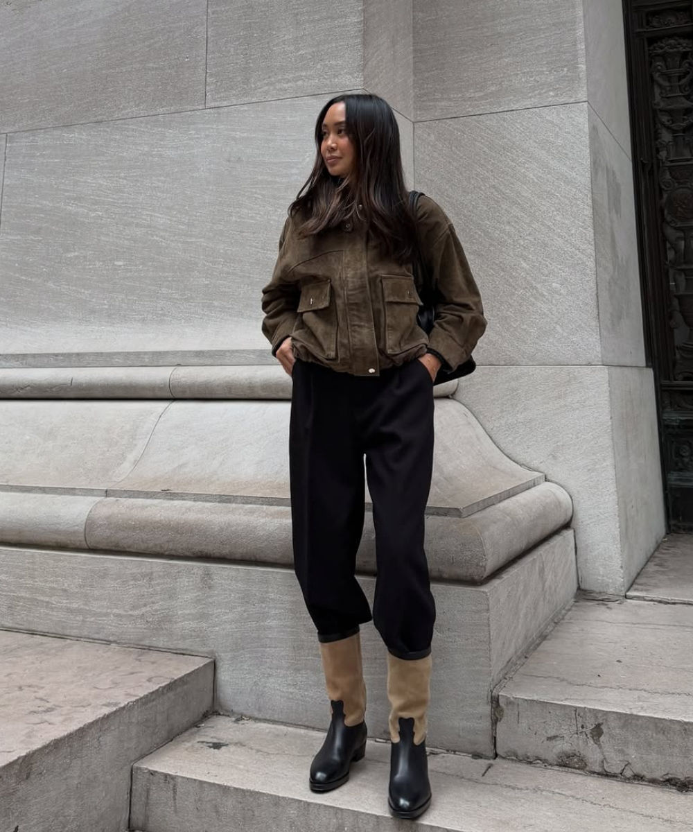 Look outono/inverno 2026: jaqueta de camurça verde-oliva, calças pretas largas e botas de cano médio bicolor. Estilo urbano e sofisticado.