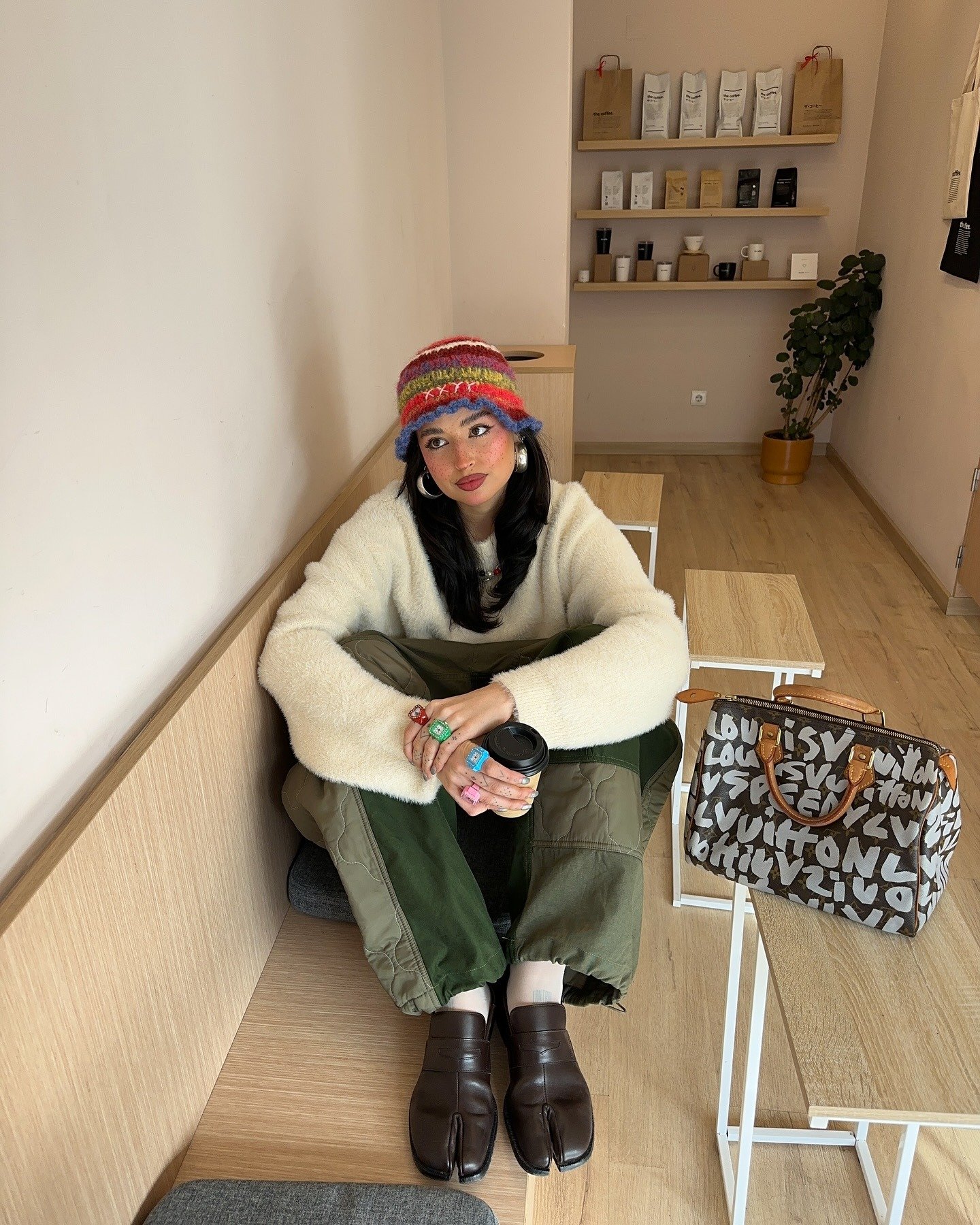 Modelo senta casualmente em banco de madeira, usando sapato tabi marrom em couro, calça cargo verde e suéter creme, com