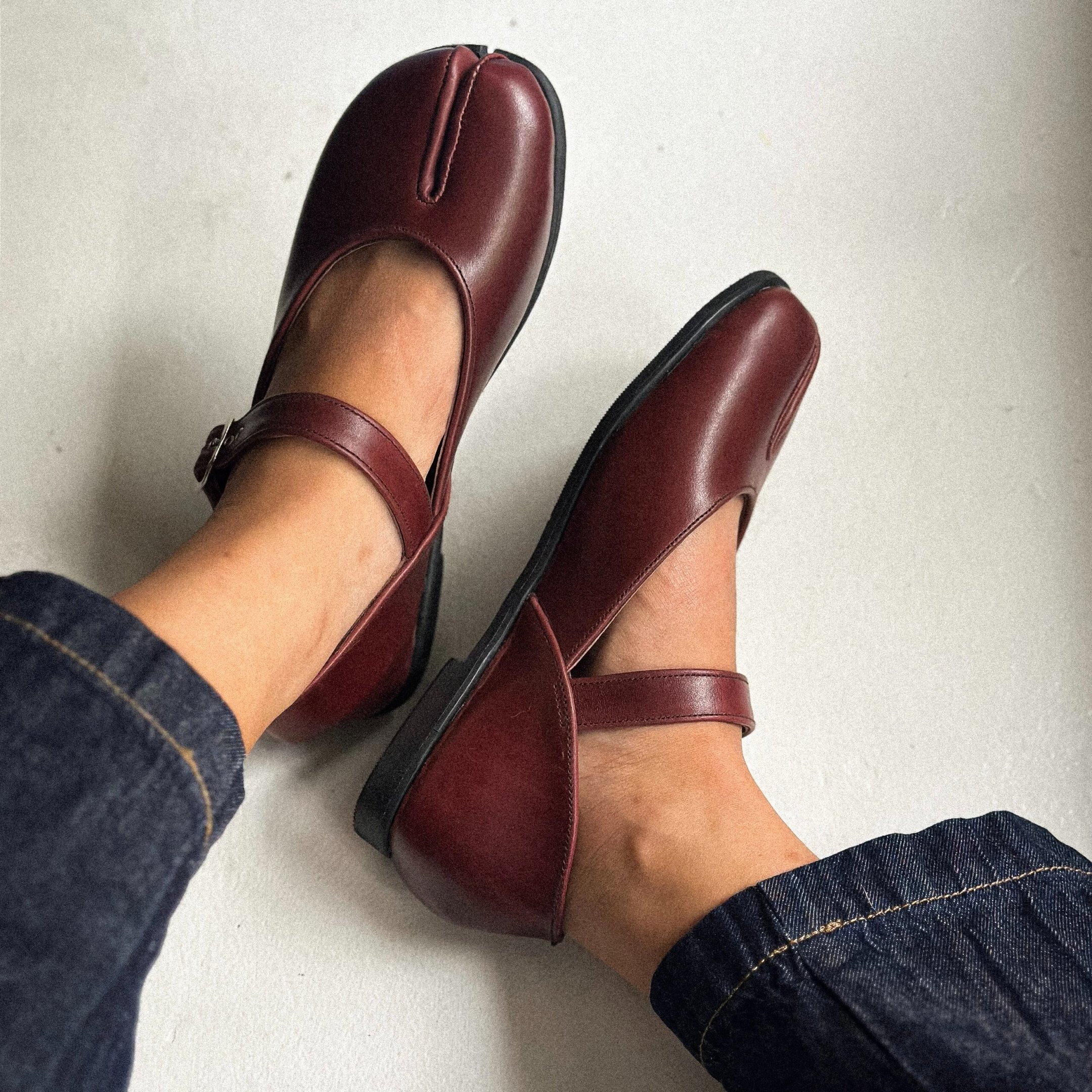 Sapato tabi em couro vinho com modelagem Mary Jane e sola preta, combinado a jeans escuro para visual moderno e confortável.