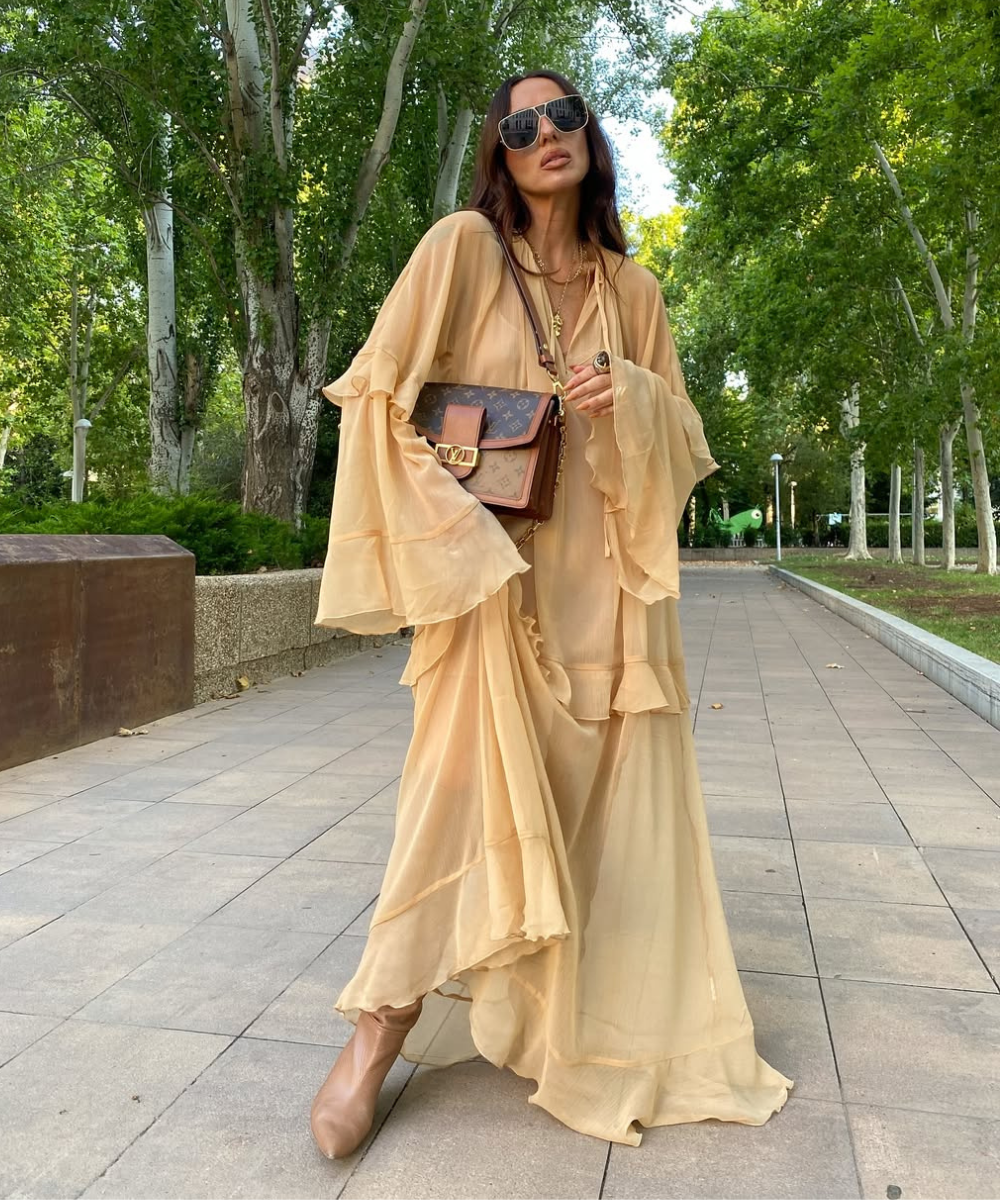 Modelo posa com vestido fluido bege de tecido leve e mangas amplas com babados, combinando bolsa marrom e clima sofisticado