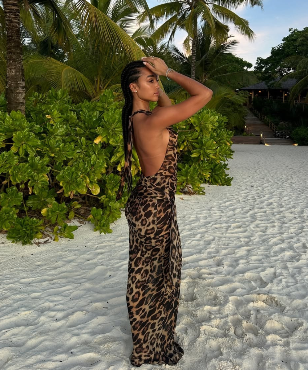 Modelo posa de perfil na praia com vestido longo de verão em tecido leve, estampa animal print e decote profundo nas costas,