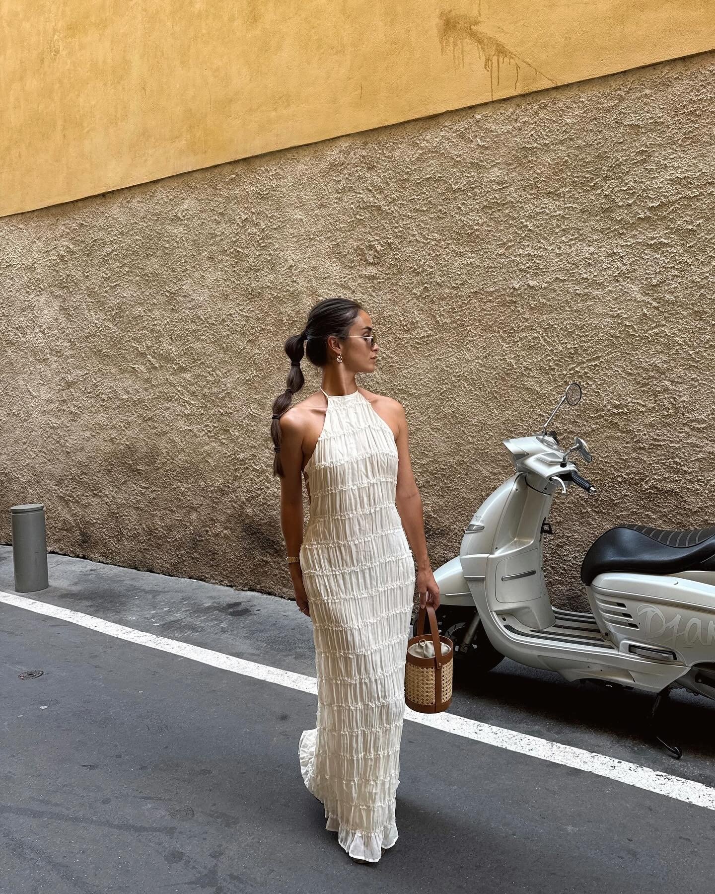Modelo posa de perfil com vestido longo branco texturizado, decote halter e bolsa de palha, em clima elegante para o final