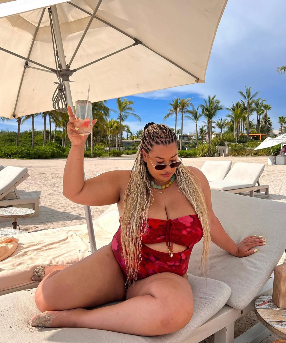 Modelos de maiô adulto em tom vermelho com detalhes em estampa, sentada em uma espreguiçadeira à beira da praia.