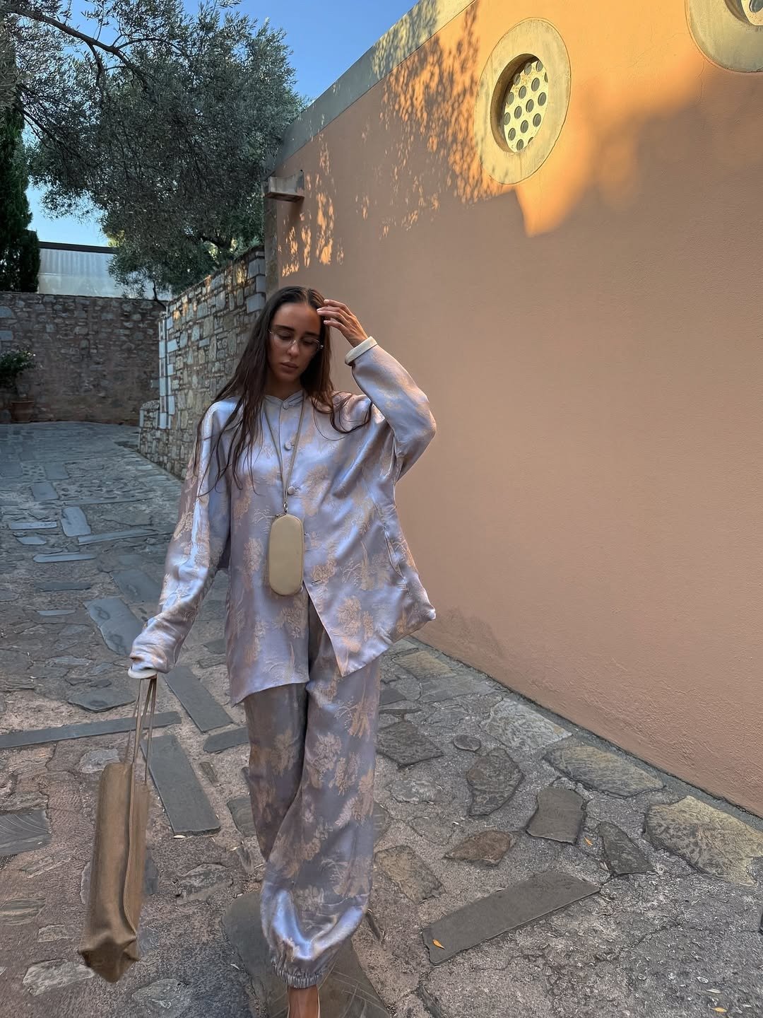 Modelo posa com colar funcional bege em cordão longo, combinando com conjunto fluido acetinado cinza com estampa floral e