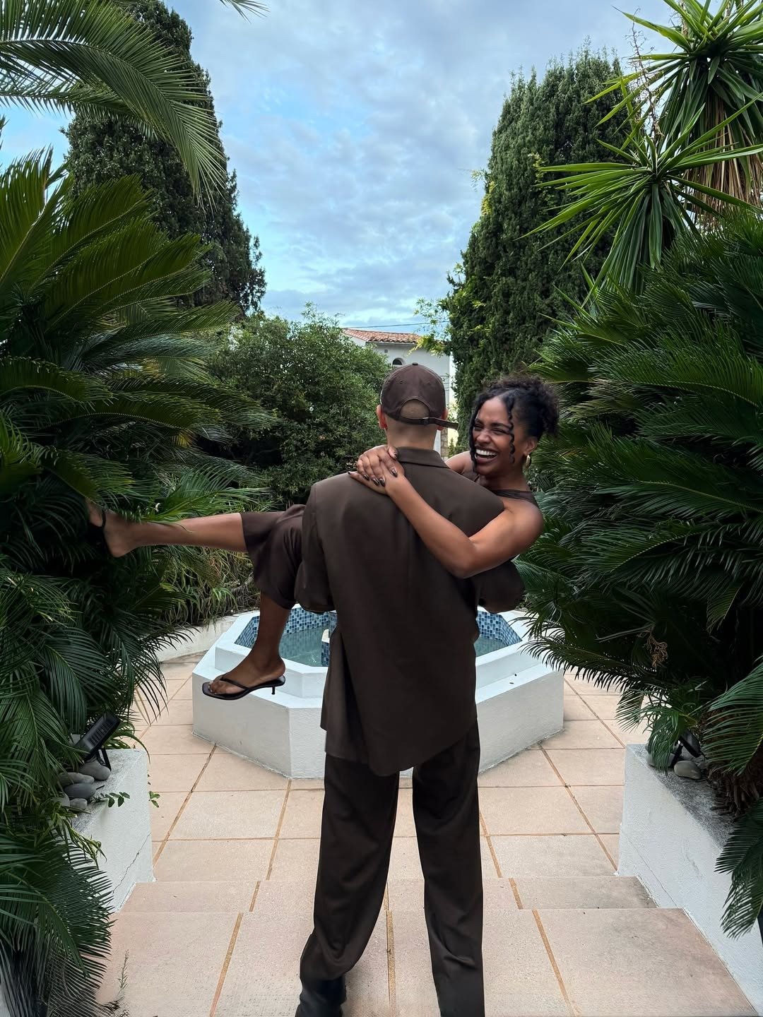 Um casal feliz se destaca em um ambiente natural, cercado por plantas exuberantes. A mulher, vestindo um vestido preto elegante, é carregada pelo ter um namorado, que usa um conjunto de roupa marrom.
