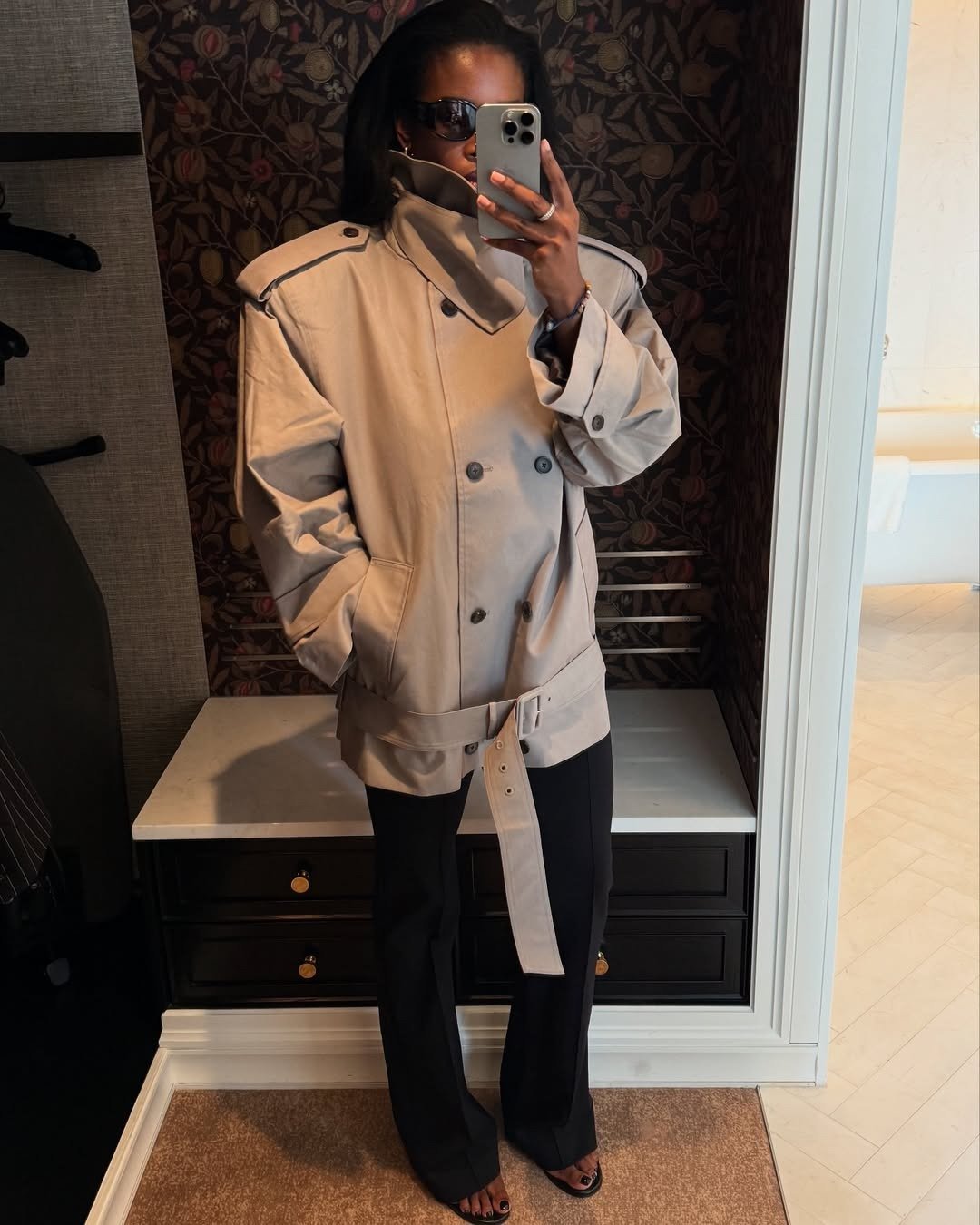 Modelo posa em jaqueta bege com gola funil estruturada, botões duplos e cinto largo, combinada a calça preta reta e sandália