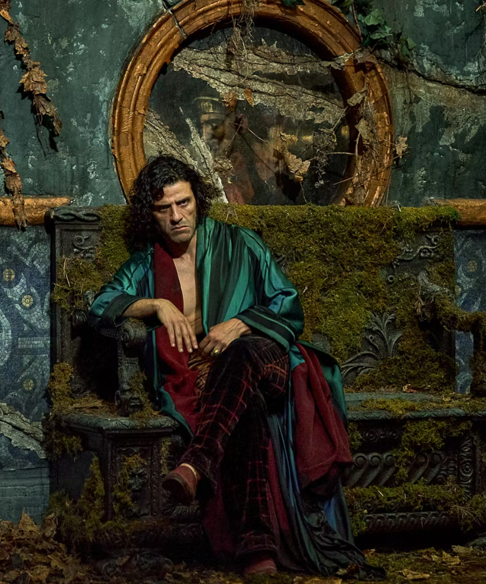 Homem sentado com expressão sombria veste roupão verde acetinado e calça xadrez vermelha, figurino de Frankenstein em