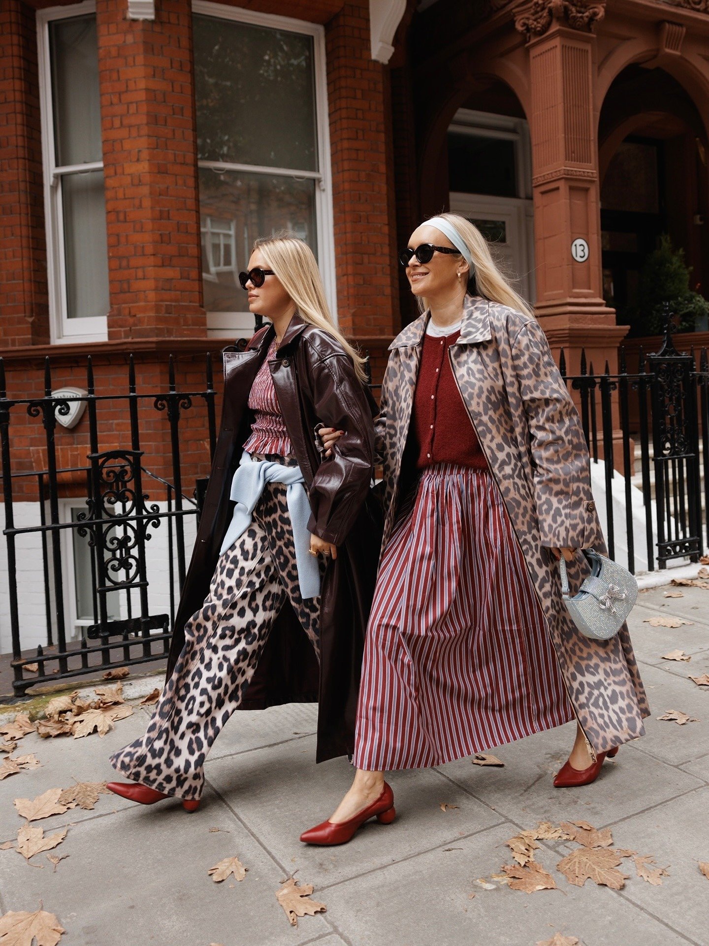 Duas mulheres caminham em uma calçada, vestindo looks estilosos e modernos. Uma usa um casaco de couro marrom, blusa de listras e calças de estampa de leopardo, enquanto a outra opta por um vestido longo listrado e um casaco de estampa de leopardo.