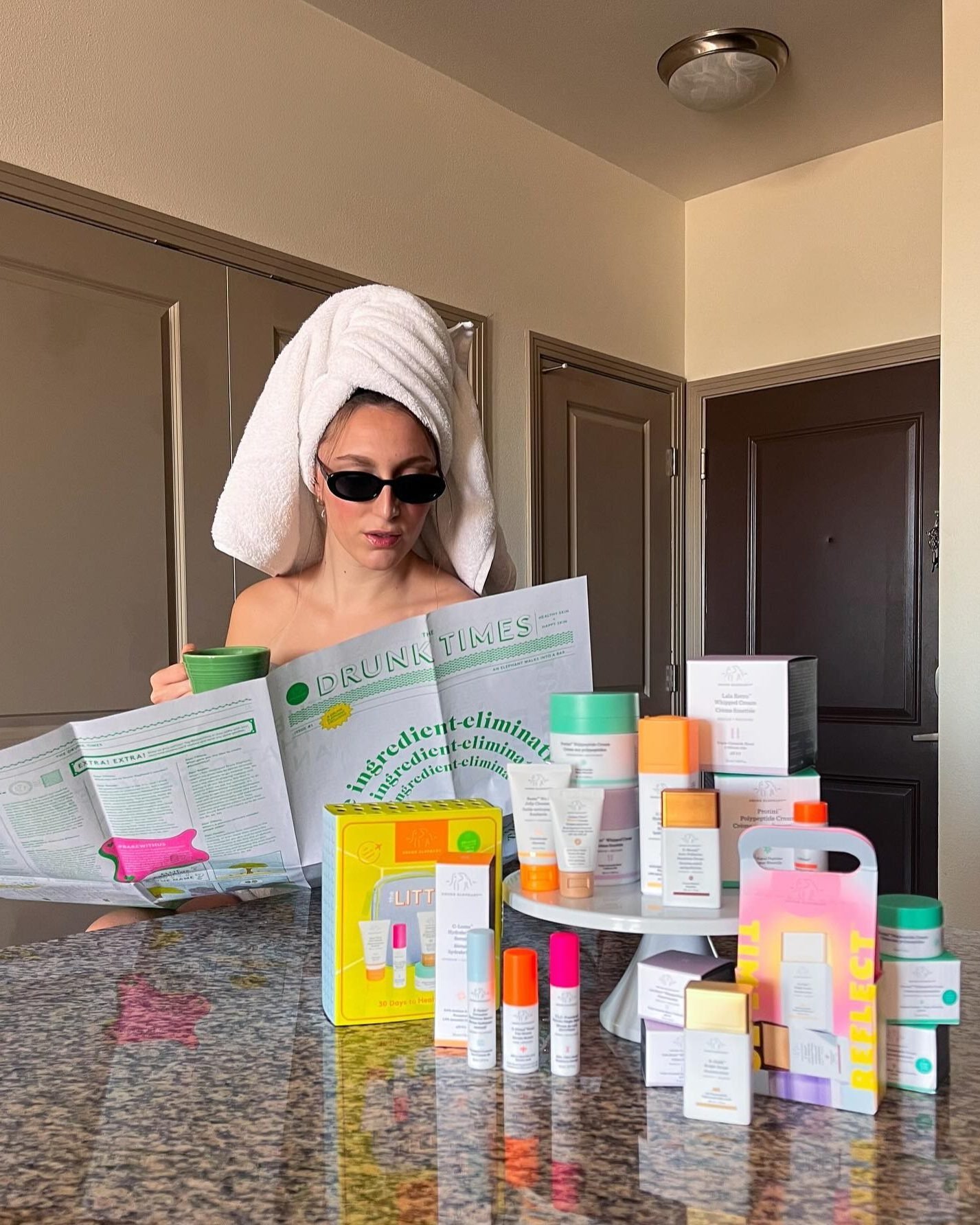 Mulher com toalha branca na cabeça e óculos escuros lê jornal segurando xícara verde, cercada por produtos coloridos para