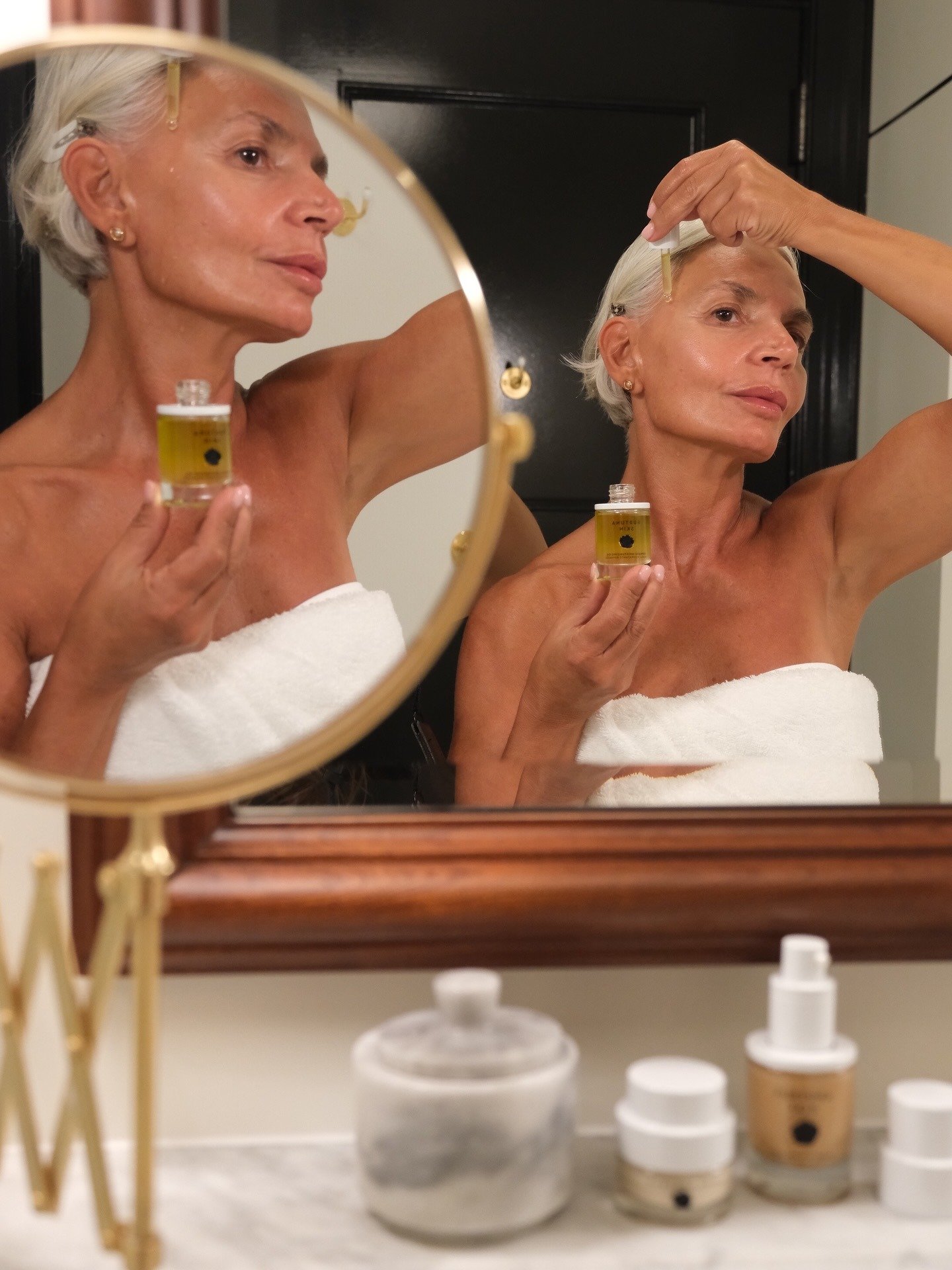 Mulher madura aplica óleo facial com delicadeza, usando toalha branca, em ambiente acolhedor para cuidar da pele ressecada.