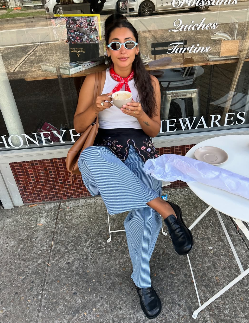 A imagem apresenta uma jovem estilosa sentada em um café, usando óculos de sol com design ousado, uma tendência forte para 2026. Ela veste uma blusa branca e calças listradas, complementadas por um lenço vermelho no pescoço, que adiciona um toque de cor.