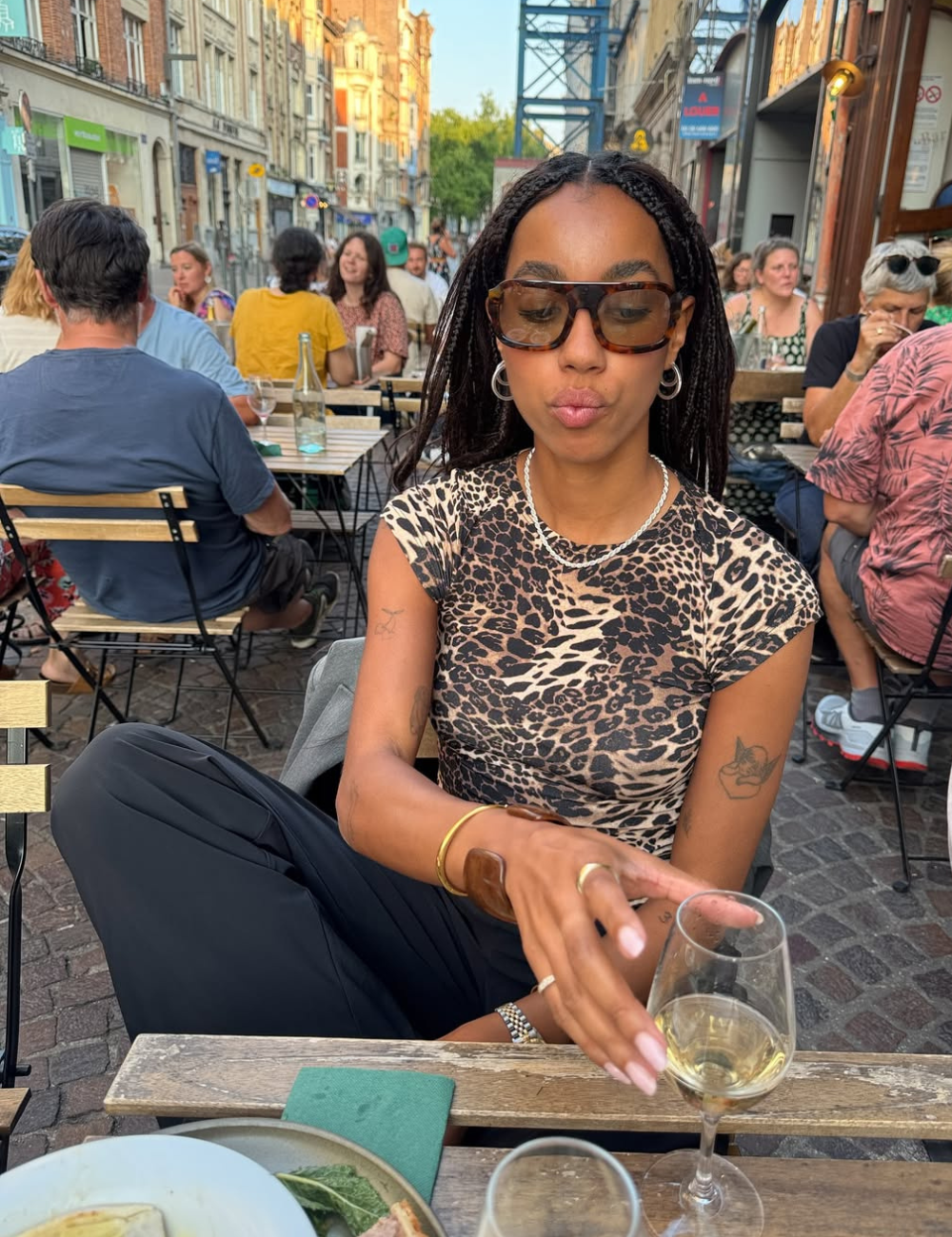 Em um ambiente ao ar livre, uma mulher exibe óculos tendência 2026, com armação grande e estilosa, alinhando-se às últimas novidades da moda. Ela veste uma blusa de estampa de leopardo, complementando sua aparência com acessórios delicados.