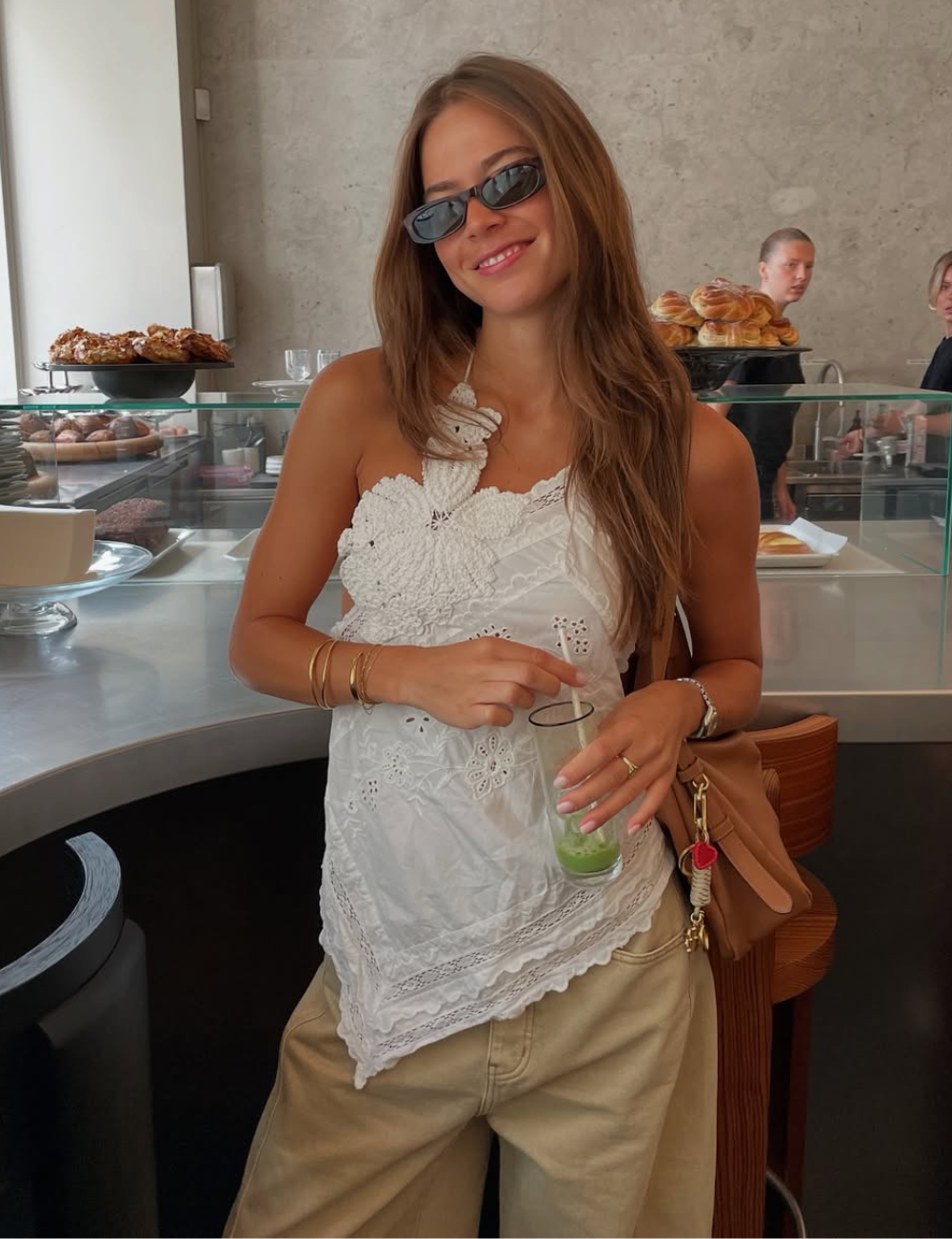 A imagem apresenta uma jovem vestindo uma blusa branca com detalhes em renda e calças largas em tom neutro, ideal para o verão. Usa um dos óculos tendência 2026, com formato quadrado e lentes escuras, adicionando um toque moderno ao look.